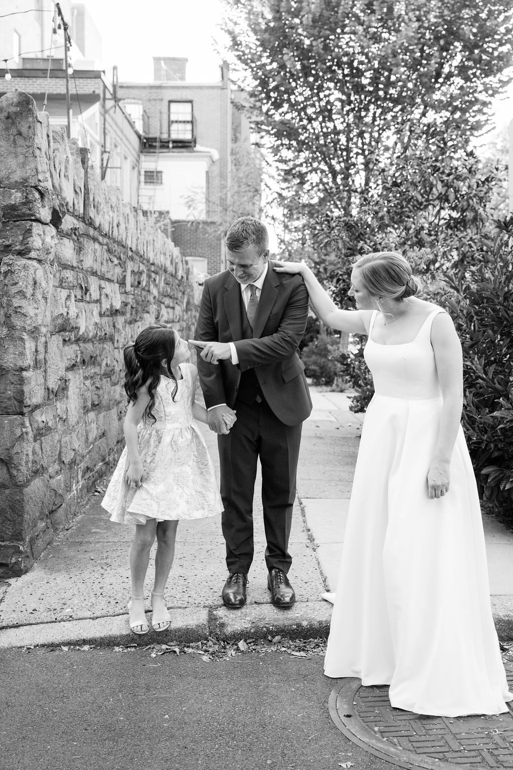 A playful wedding moment captured on a sidewalk beside a stone wall.