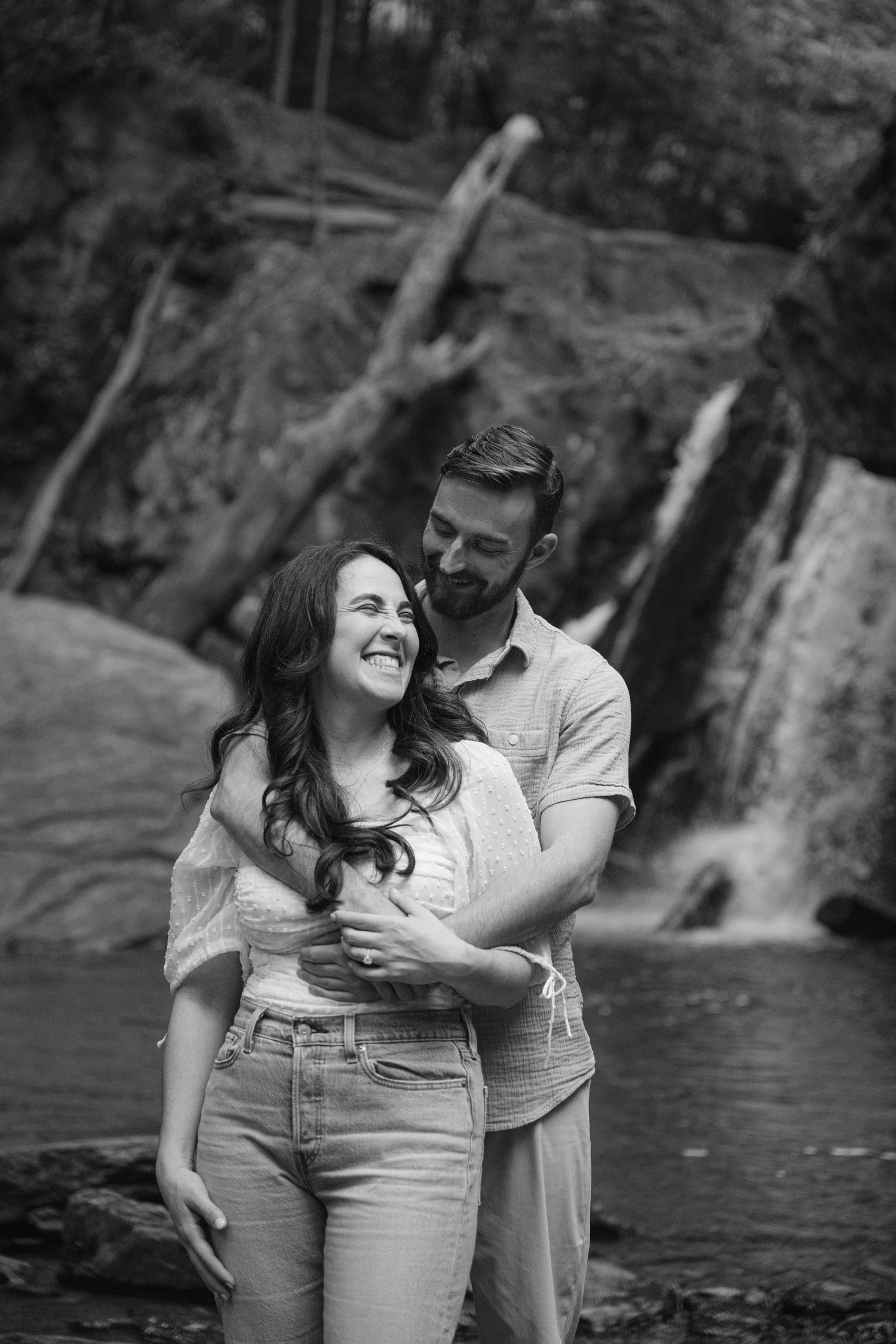 A joyful couple laughs together by a scenic waterfall in black and white.
