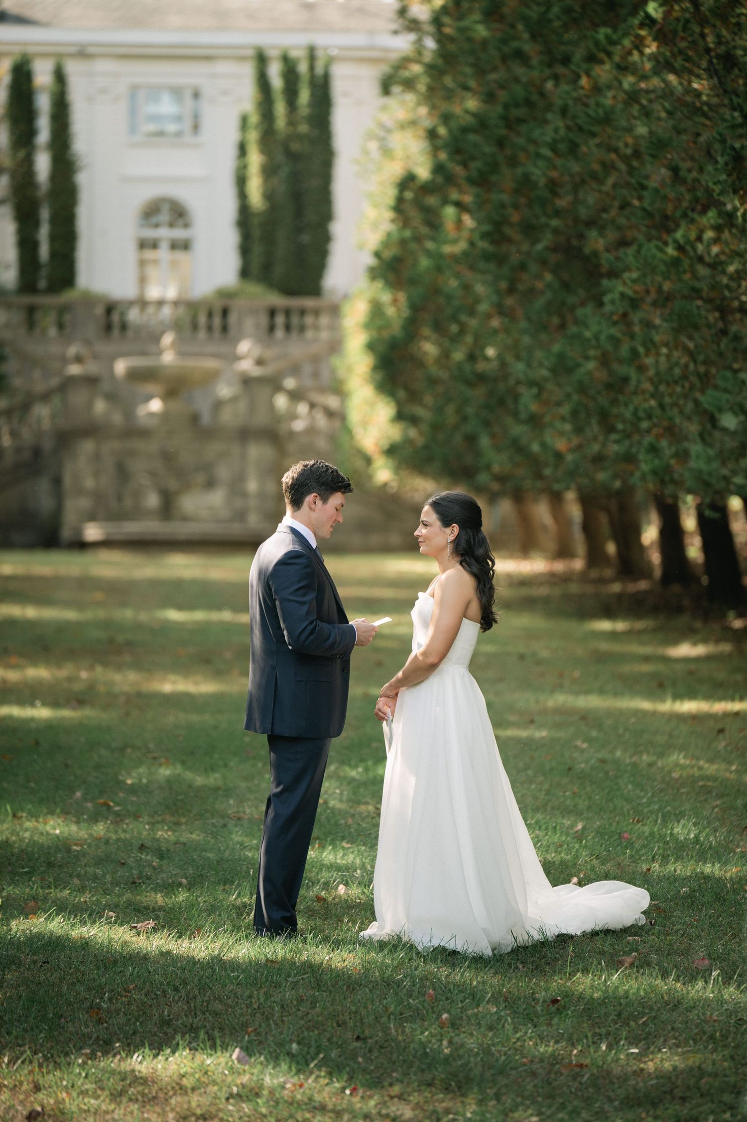 A bride and groom share a tender first look moment on a lush green lawn beside an elegant estate.