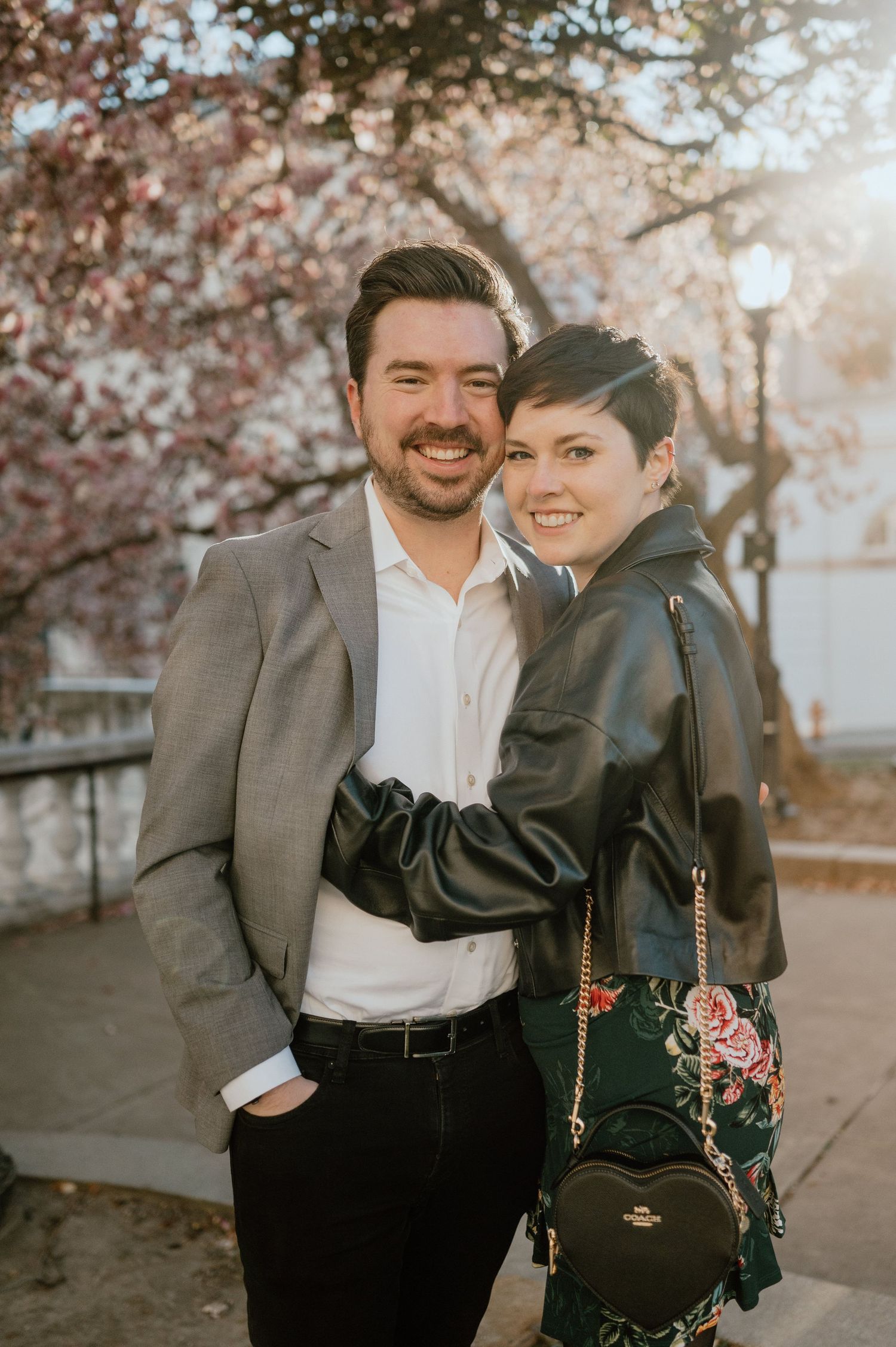 A smiling couple poses outdoors near cherry blossoms, man in gray blazer and woman in leather jacket with floral skirt.