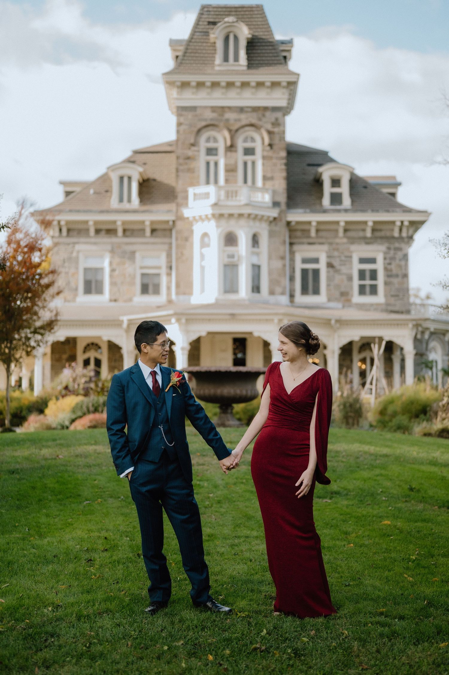 Couple in formal attire holding hands on lawn before a grand Victorian stone mansion during autumn.