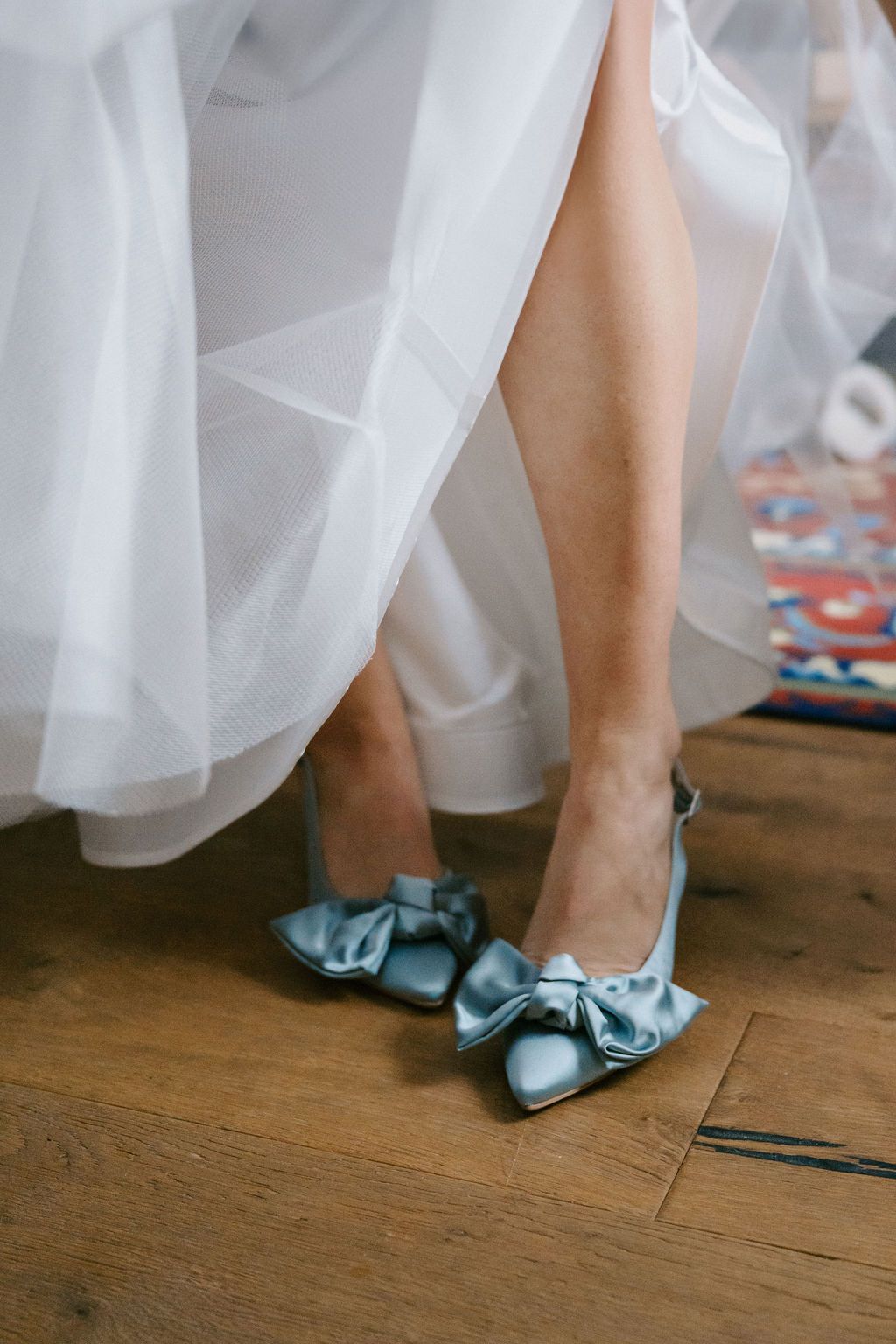 Light blue bridal shoes with bows peek out from under a flowing white wedding dress on hardwood floor.