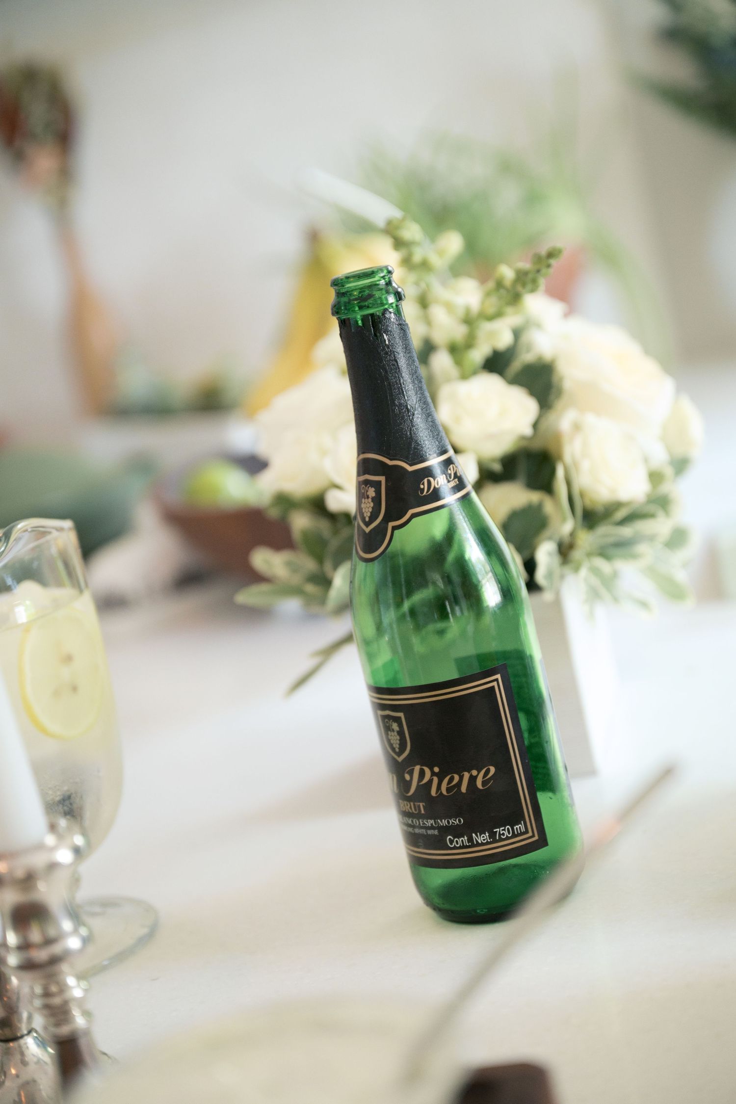 Green champagne bottles with elegant black labels sit on white tablecloth alongside delicate floral arrangements.