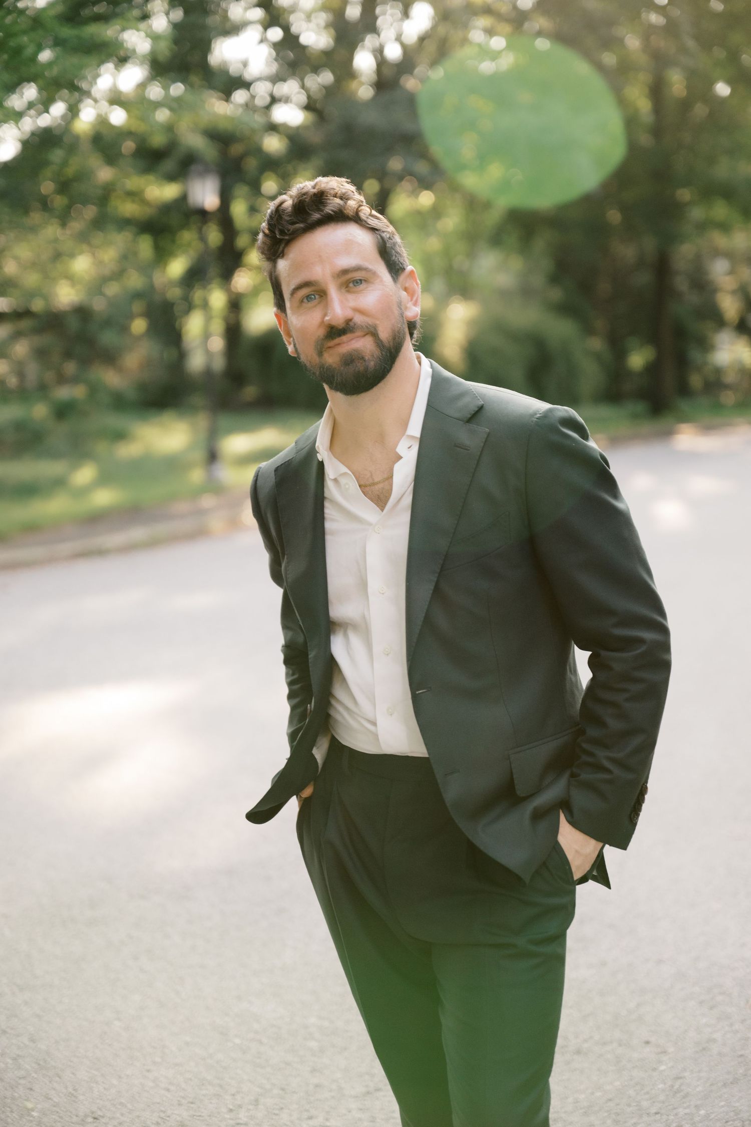 A groom poses casually in a dark suit and white shirt against a sunlit outdoor backdrop.