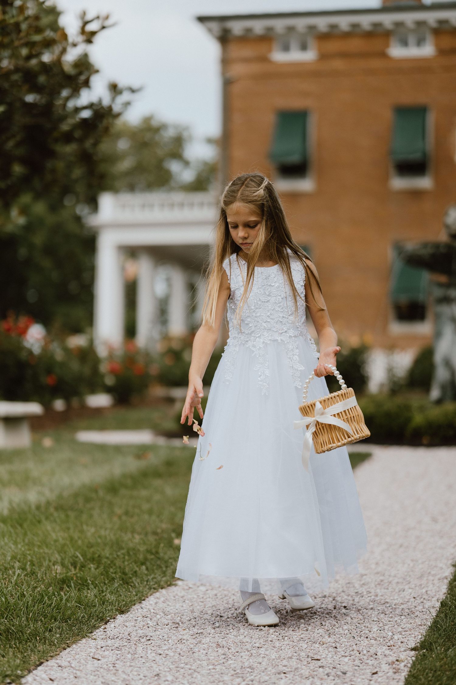 A young attendant in a white dress walks down a garden path carrying a basket.