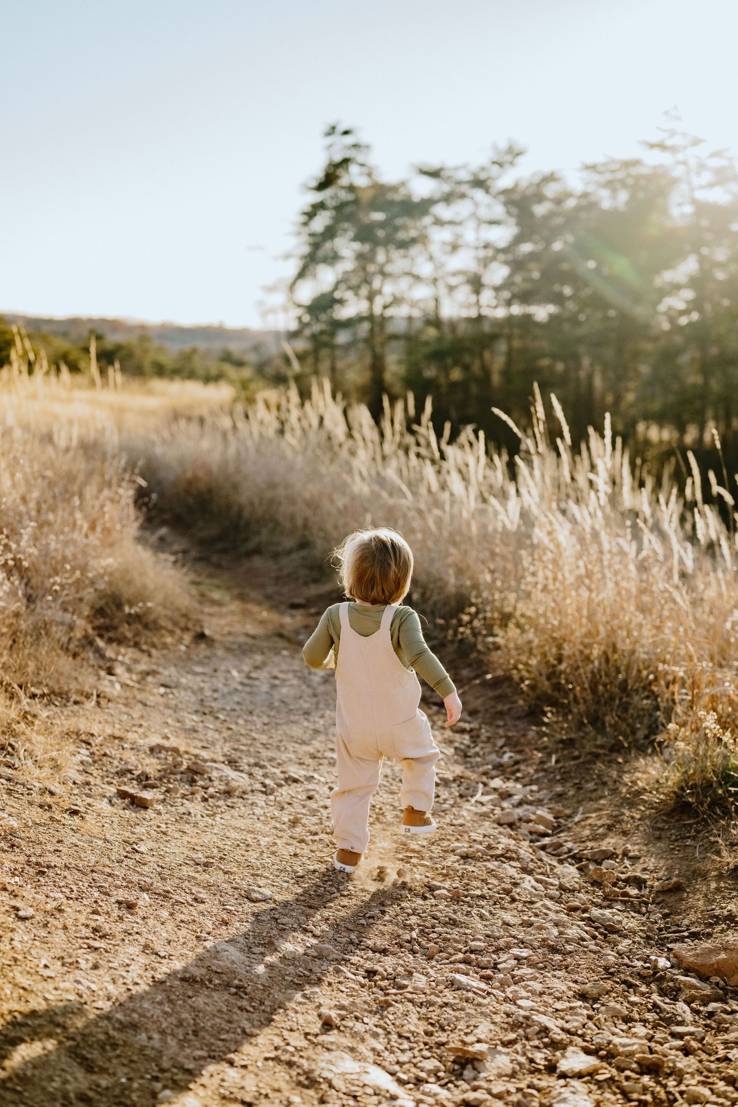 A small child in overalls runs along a sunlit dirt path through tall grass.