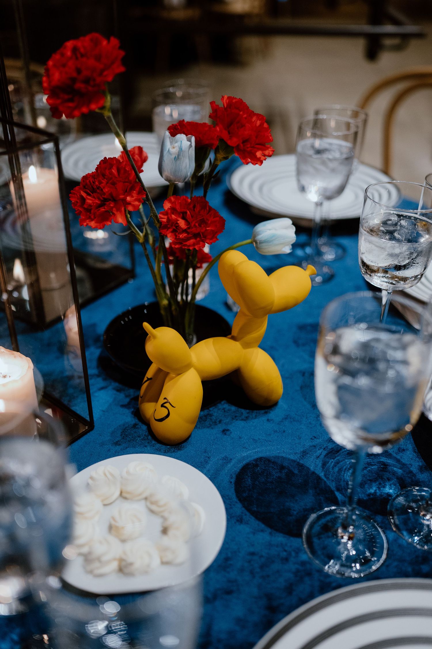 Table decor featuring red carnations, yellow balloon animal, and formal place settings on blue velvet.