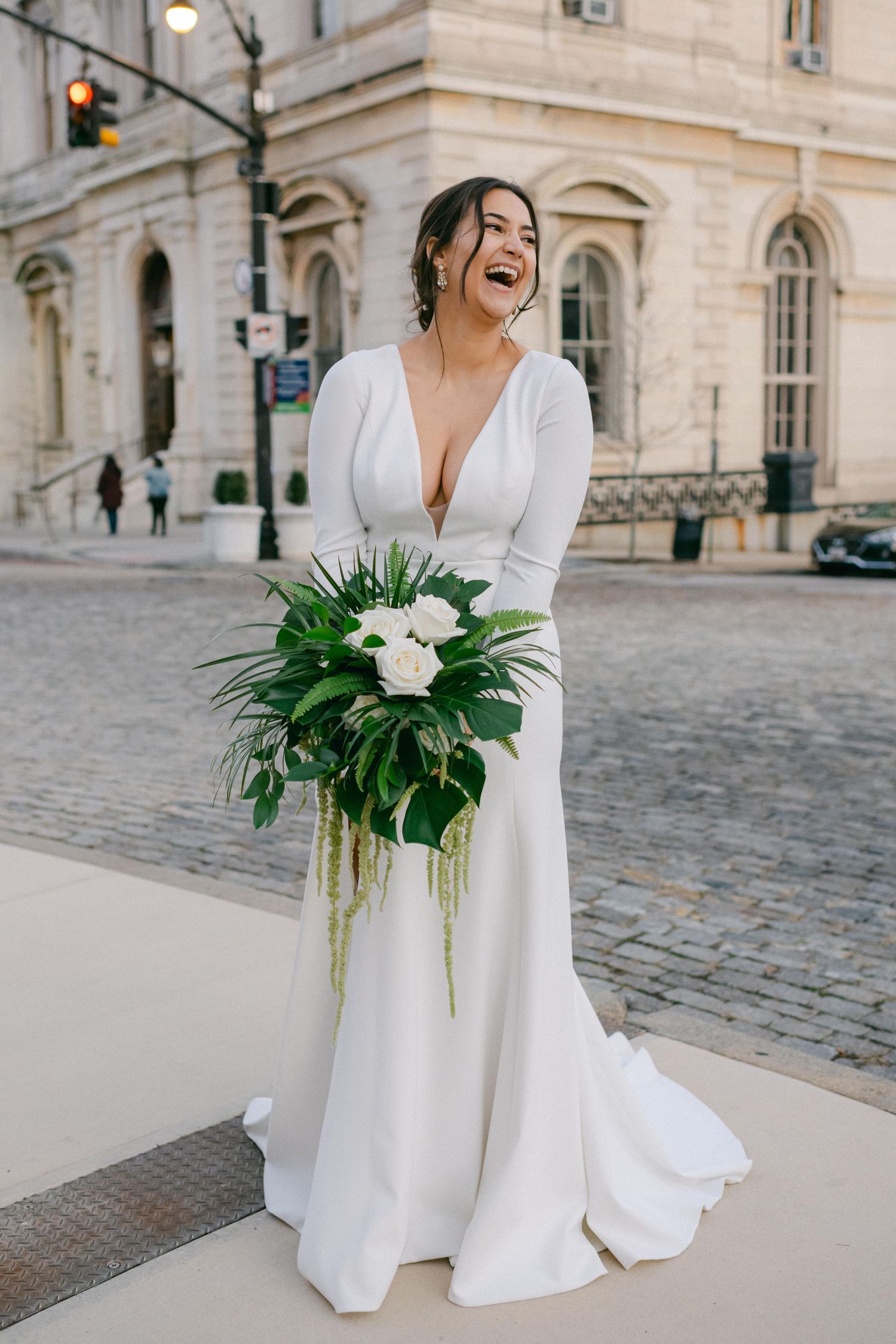 Joyful bride in sleek white wedding dress holds modern tropical bouquet outside historic building.