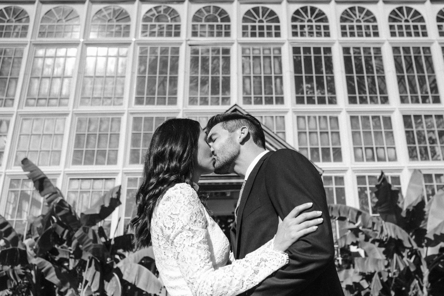 A black and white photo of newlyweds kissing in front of an ornate glass conservatory wall.