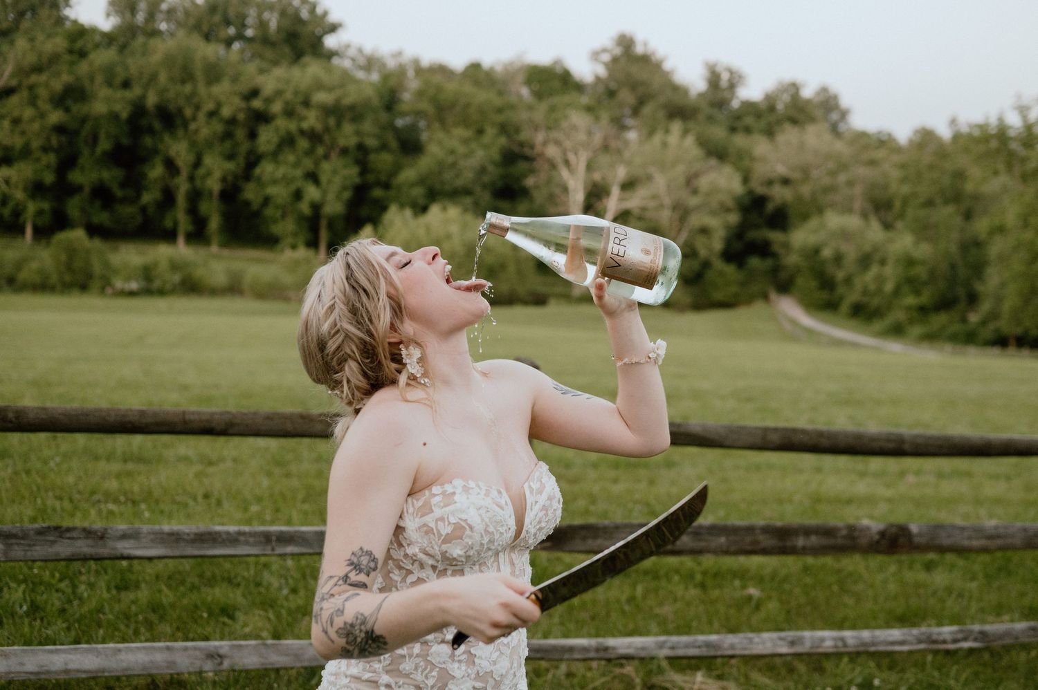 A person in a wedding dress drinks from a bottle while standing by a wooden fence.