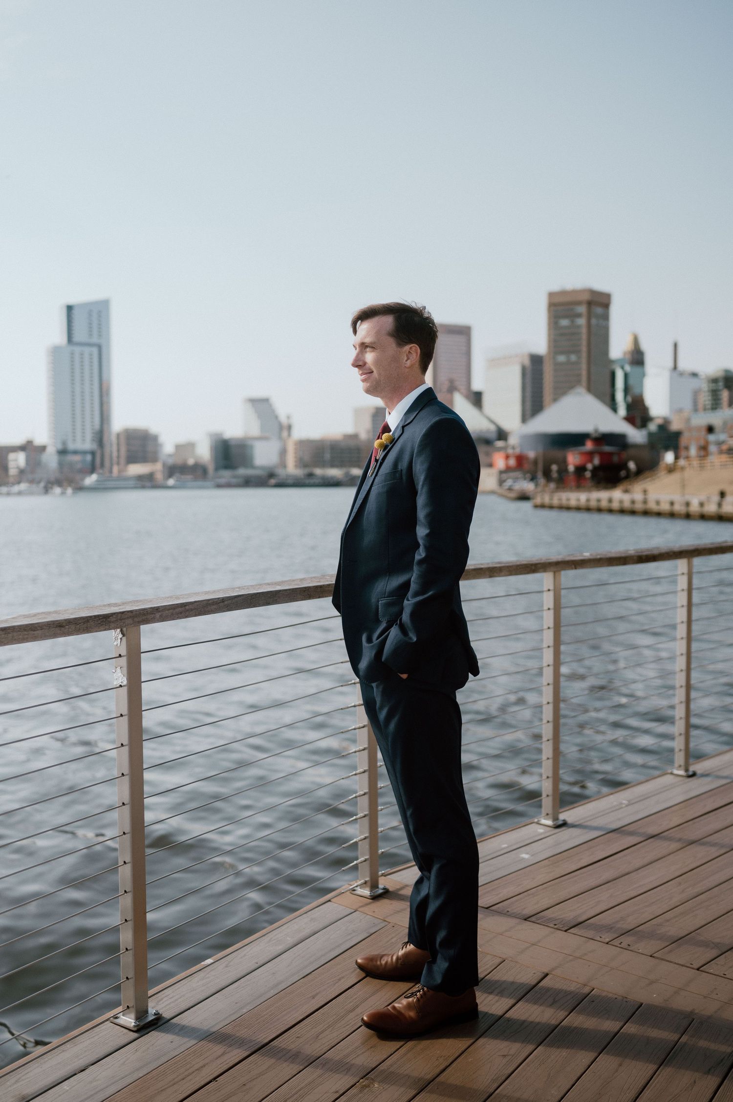 Groom in dark suit stands on waterfront deck with city skyline in background.