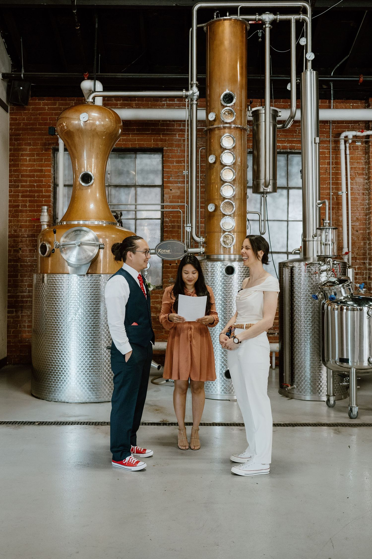 Three people standing in front of copper distillery equipment during a ceremony.