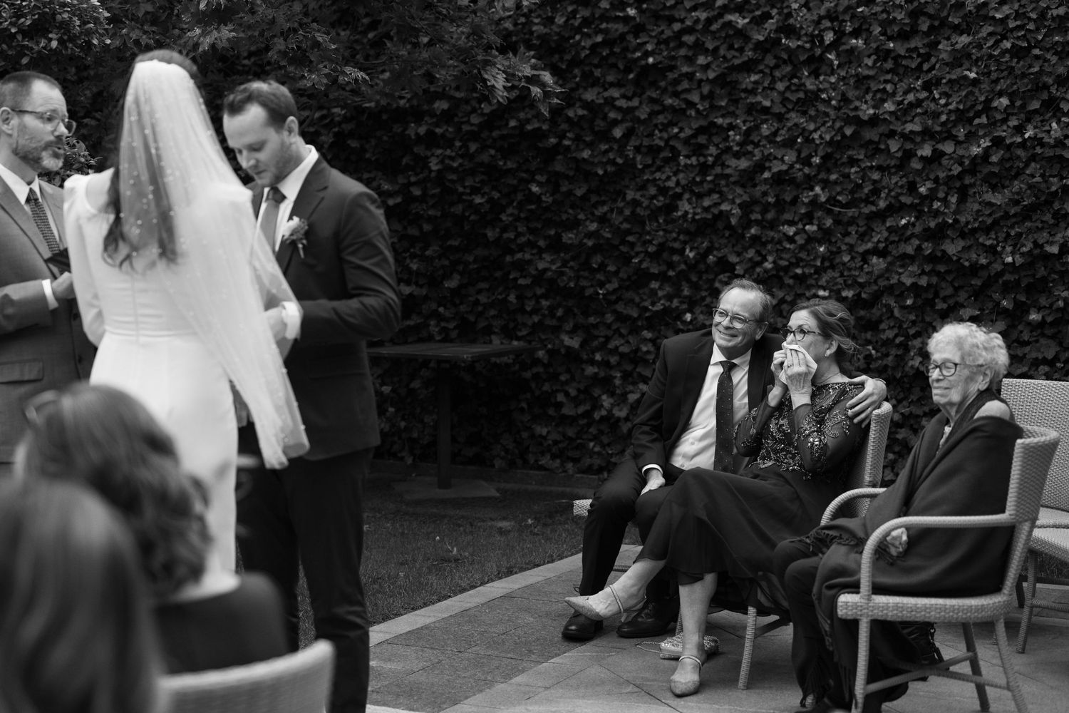 Black and white photograph captures a tender wedding ceremony moment against ivy-covered walls.