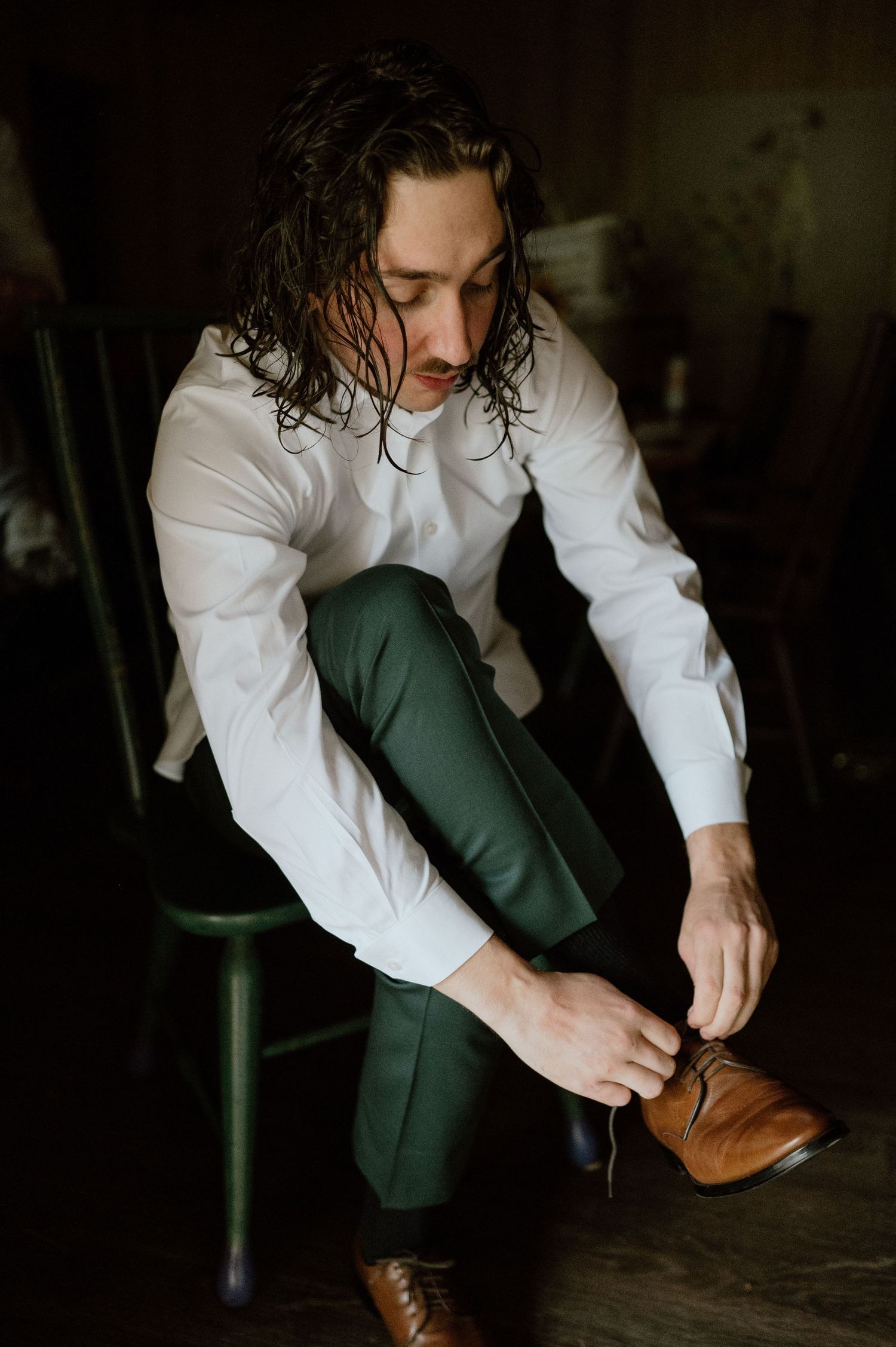 Person in formal attire putting on brown leather shoes while seated on a green chair.