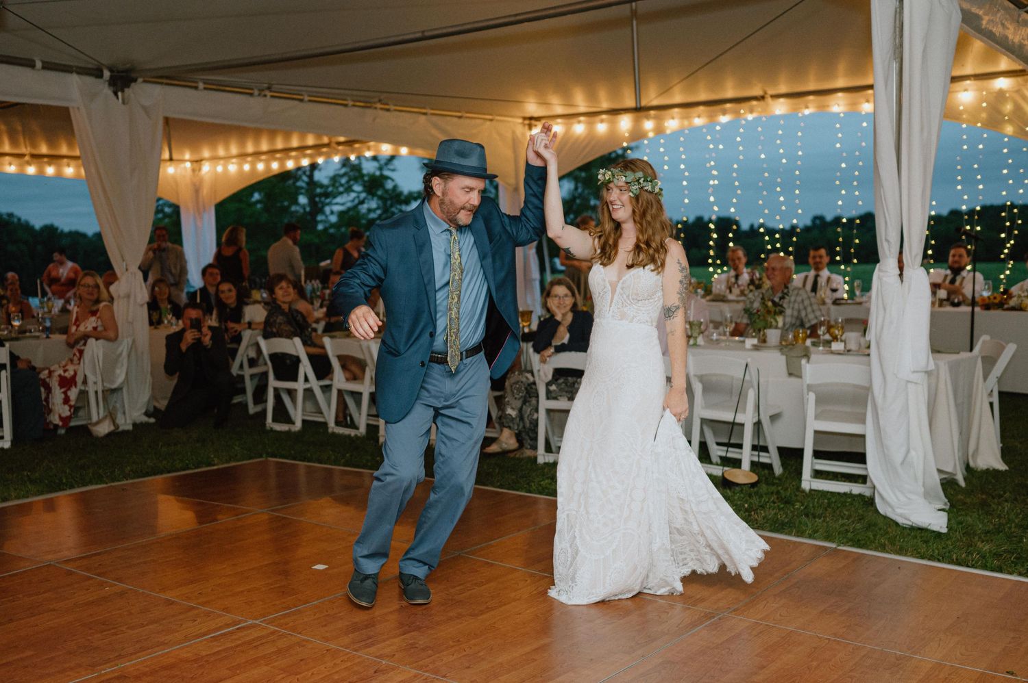Wedding couple dancing on wood floor under tent with string lights at dusk.
