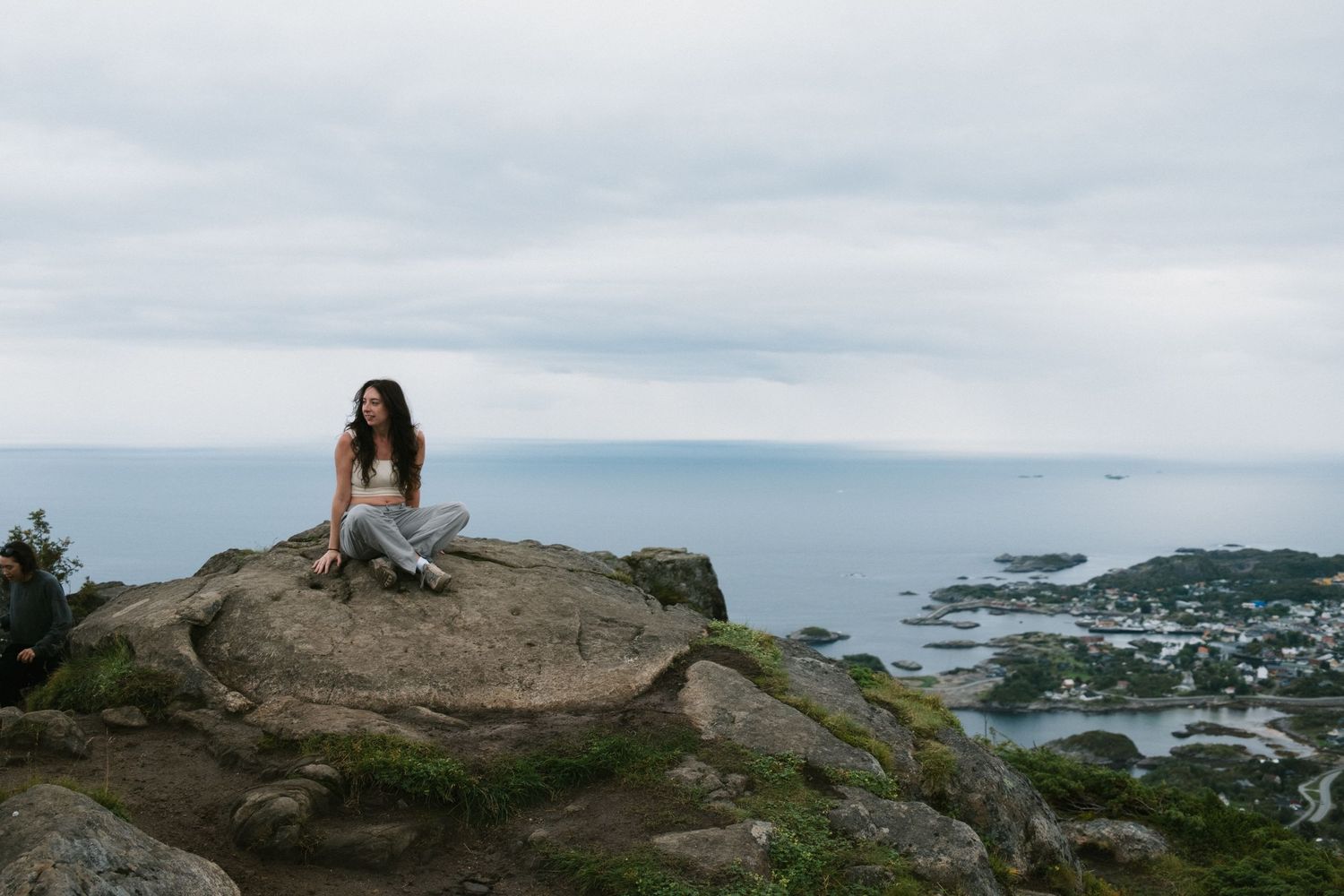Figure sits atop a rocky outcrop overlooking a dramatic coastal landscape with islands in the distance.