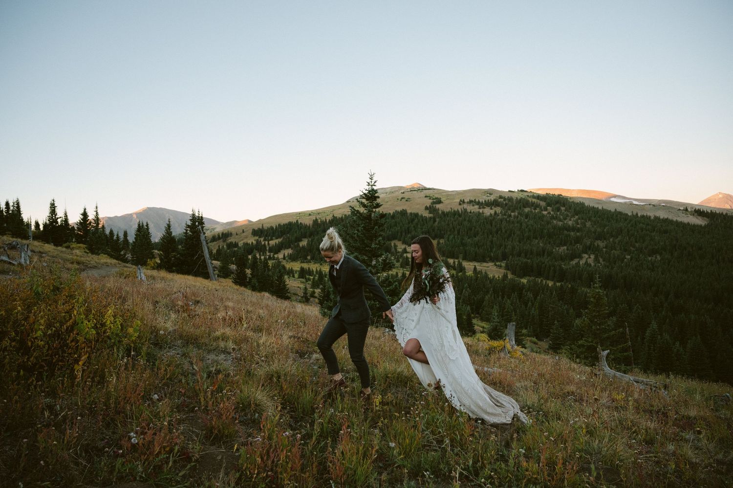A couple in wedding attire runs along a scenic mountain hillside at sunset.