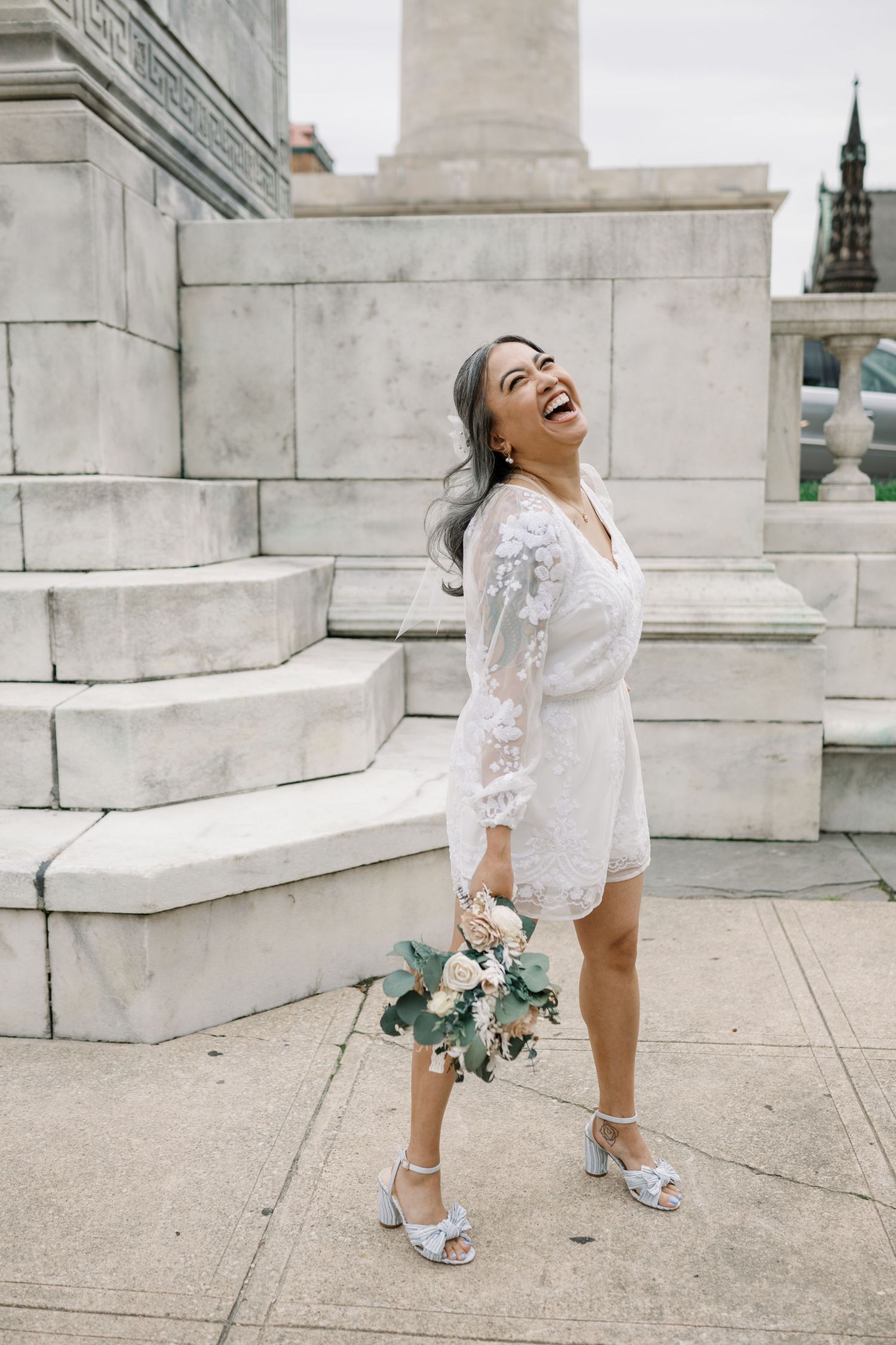 A bride in a short white lace dress laughs while holding a bouquet on marble steps.