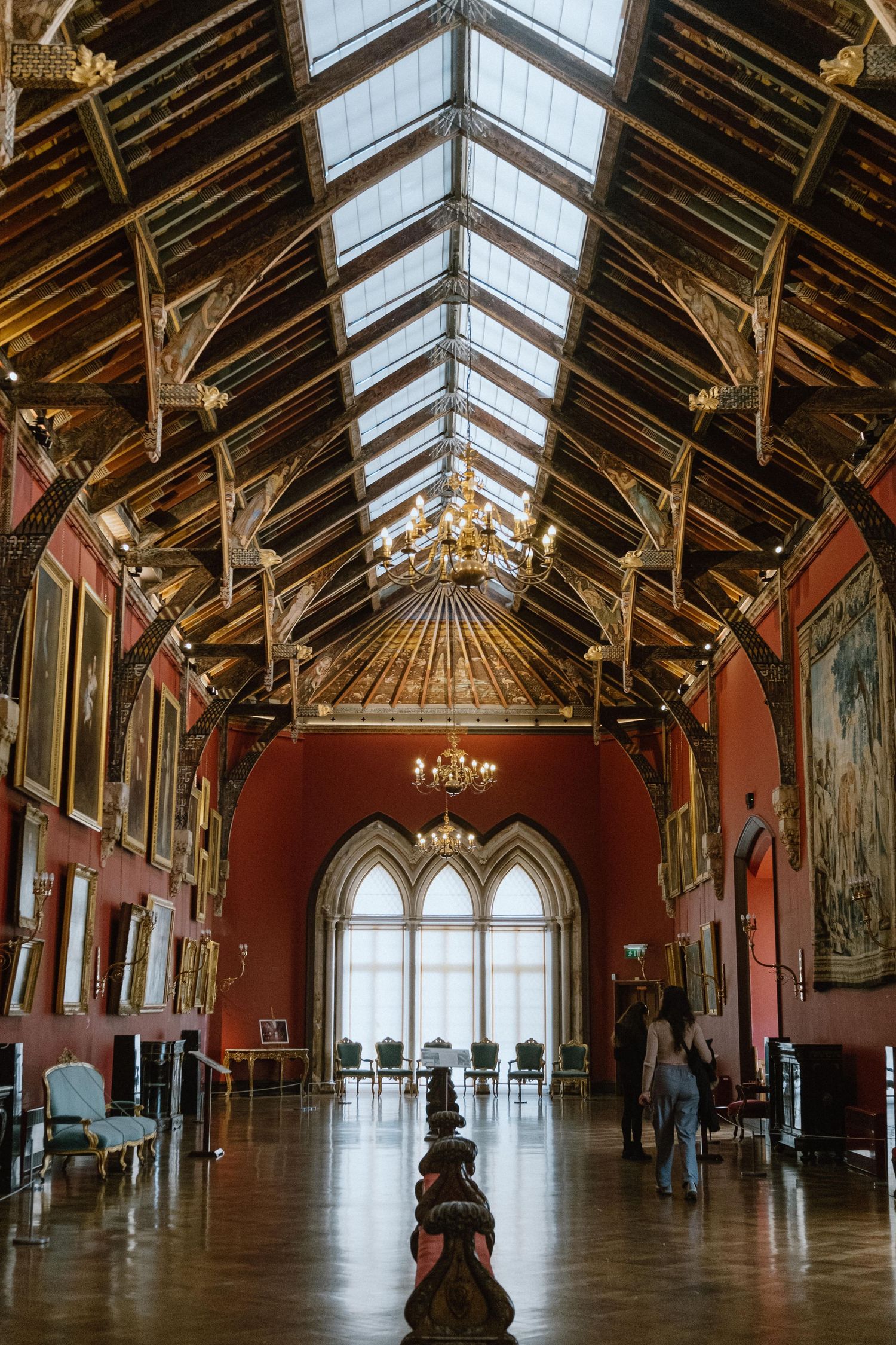 Grand medieval hall with vaulted glass ceiling, red walls, and gothic windows.