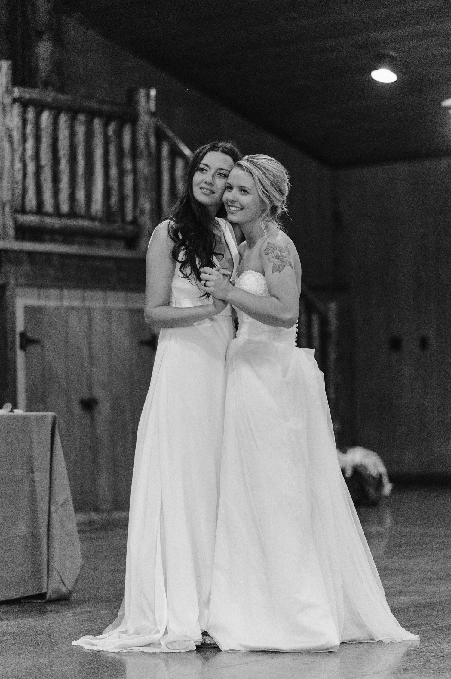 A black and white candid moment of two people in flowing white gowns embracing during a celebration.