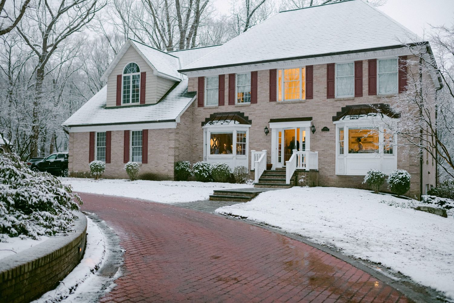 Large brick colonial style house with snow covered driveway and glowing windows at dusk.