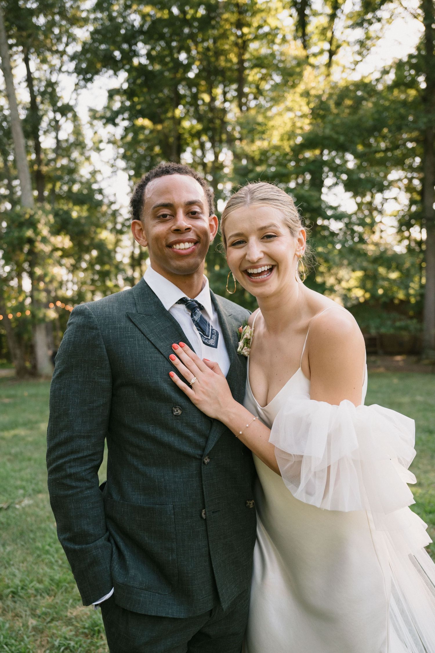 A joyful wedding couple poses together outdoors in formal attire with natural backlit scenery.