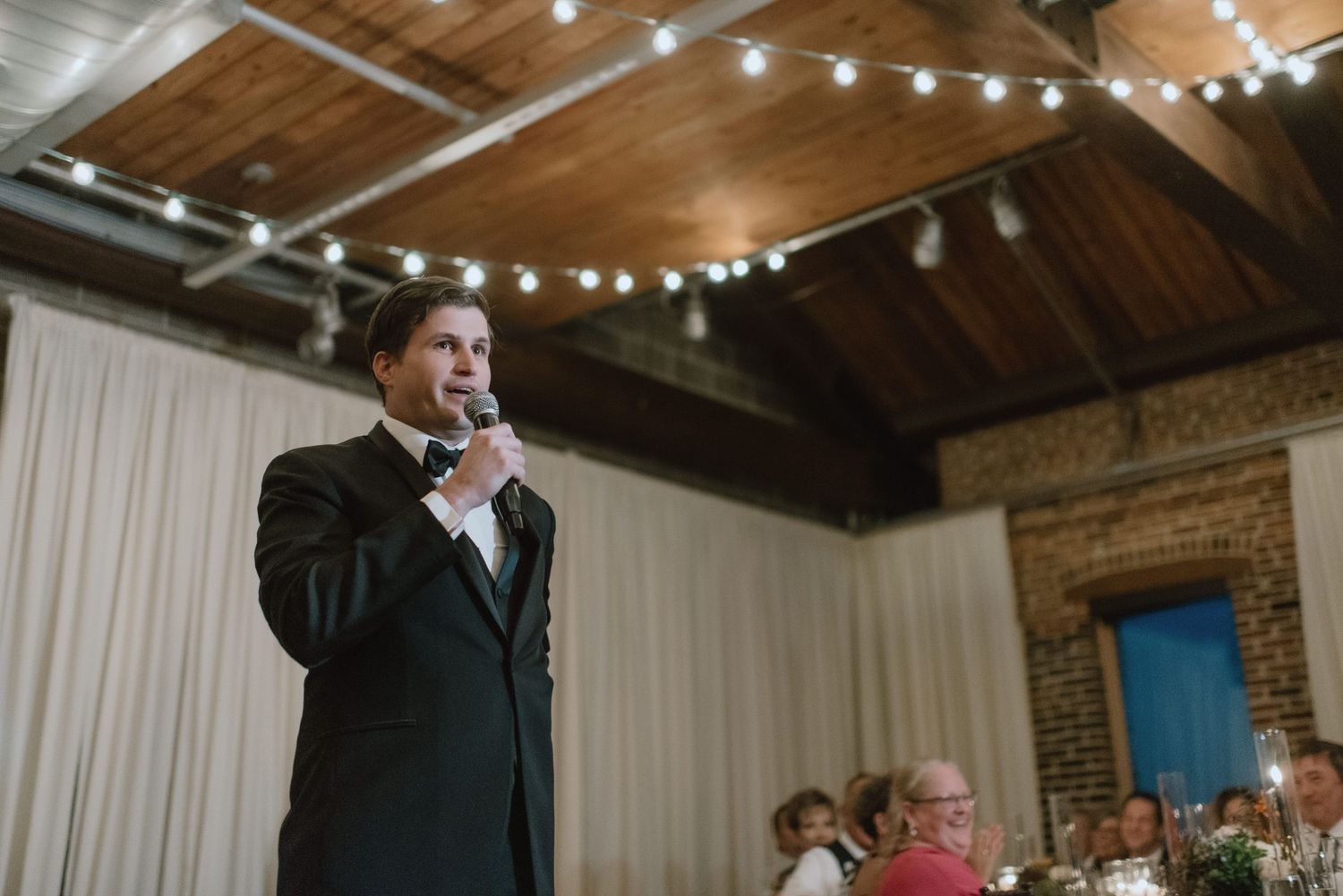A speaker delivers a toast in a rustic venue with string lights and exposed wooden beams overhead.