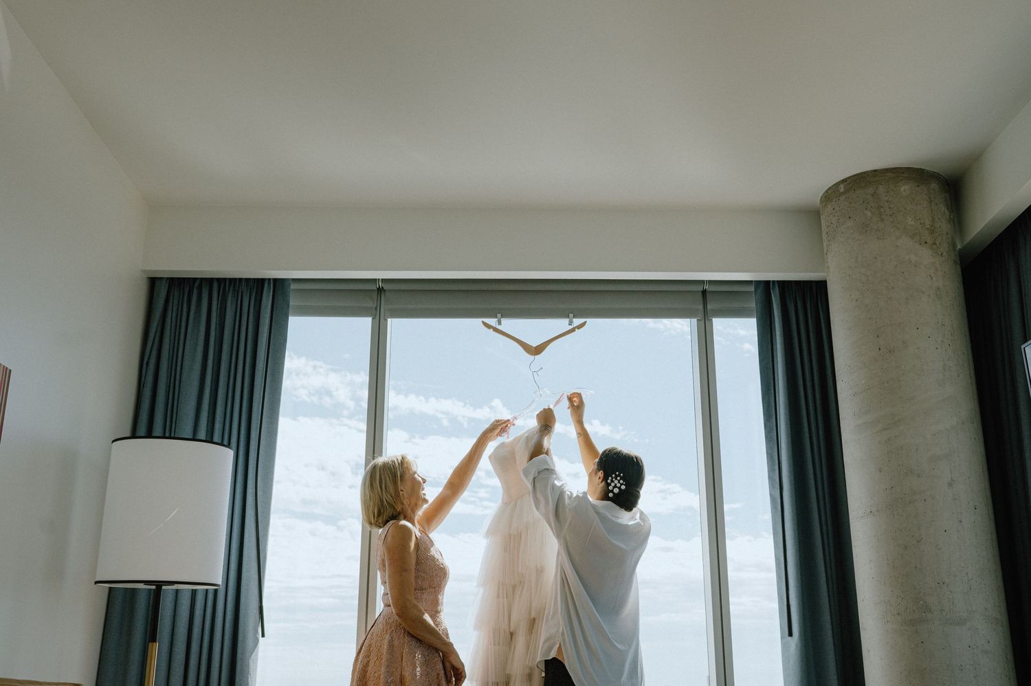 Wedding party holding up a white dress against large windows in a hotel room.