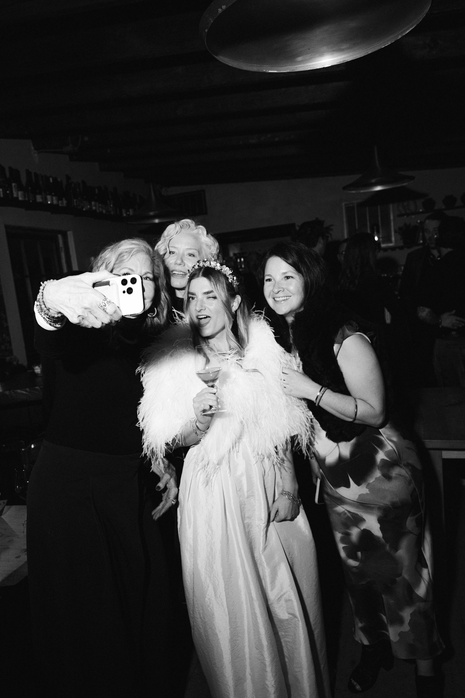 Wedding celebration with guests taking photos in a dimly lit room featuring a bride in a white dress with feather boa.