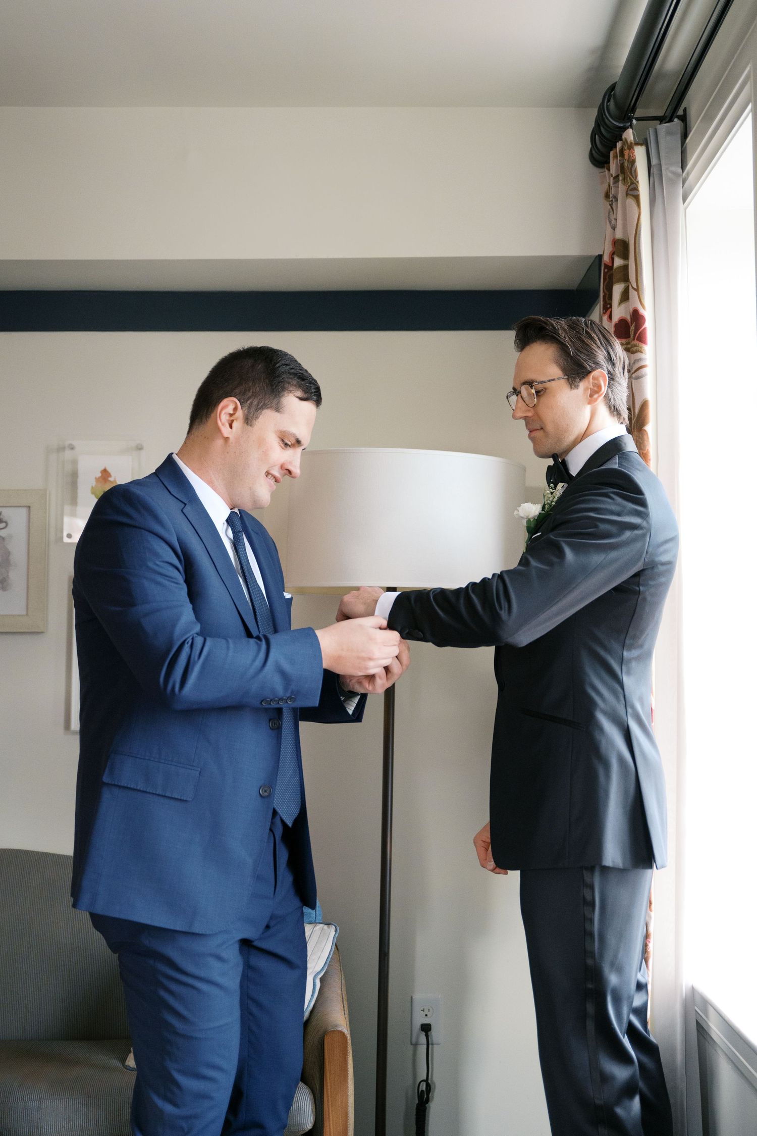 Two men in suits help each other get ready in a room with natural light from a window.
