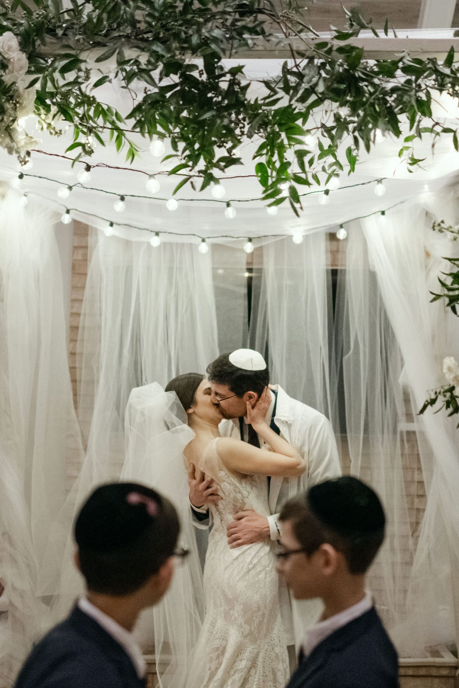Romantic wedding kiss beneath decorative arch adorned with greenery and string lights.