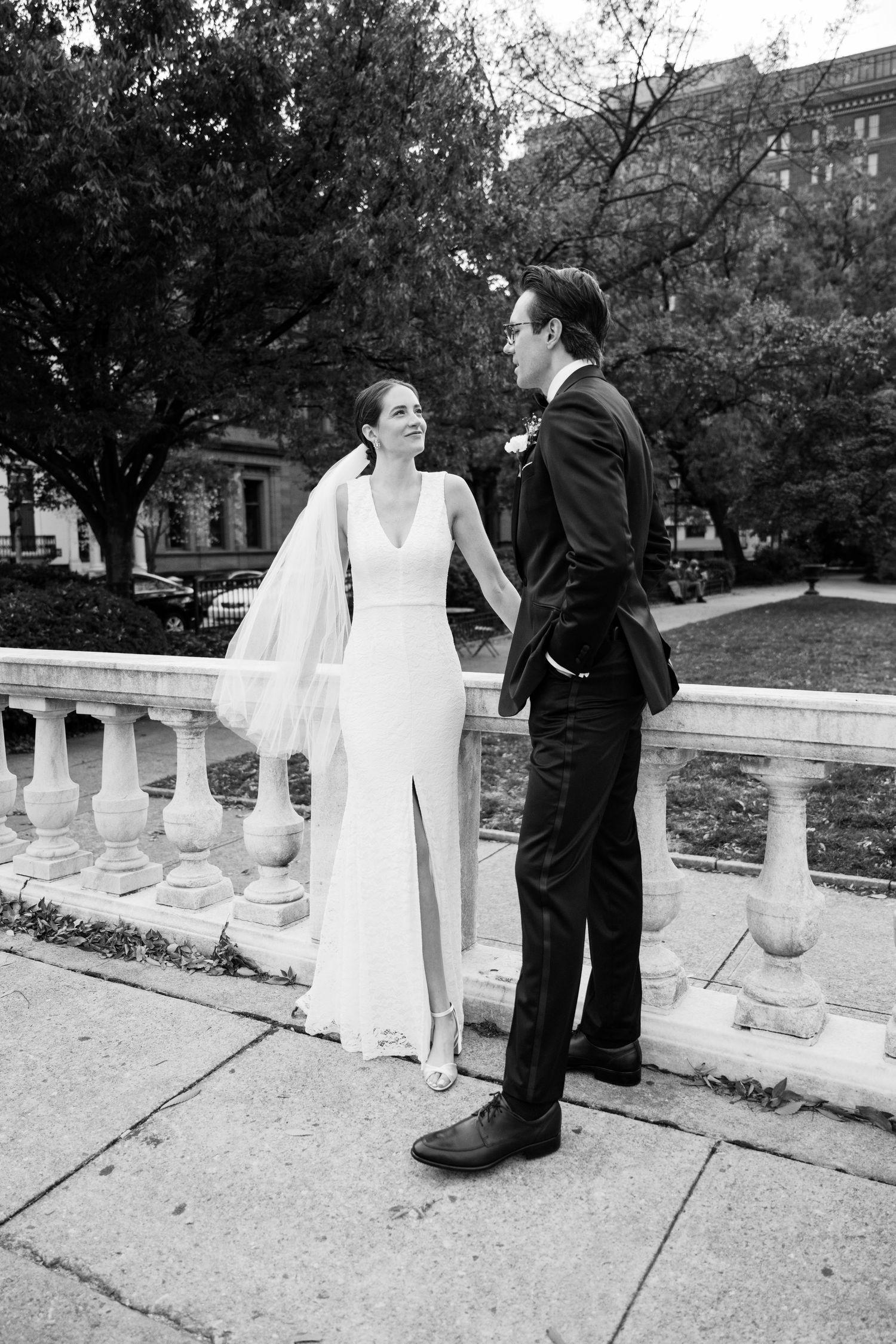 A bride and groom share a moment on a stone balustrade in an urban garden setting.