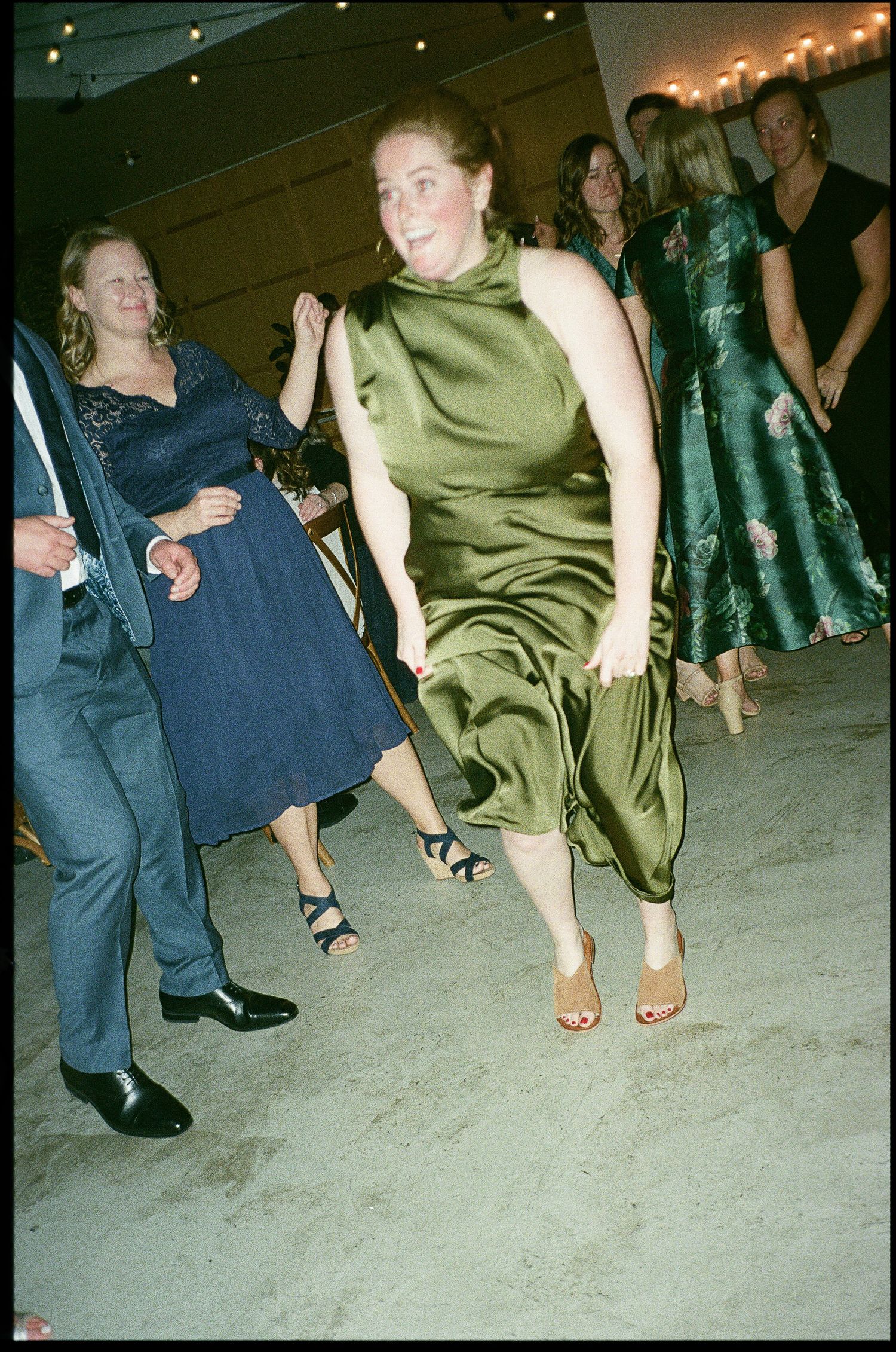 People dancing enthusiastically at a formal event, with one wearing an olive green satin dress jumping mid-dance.