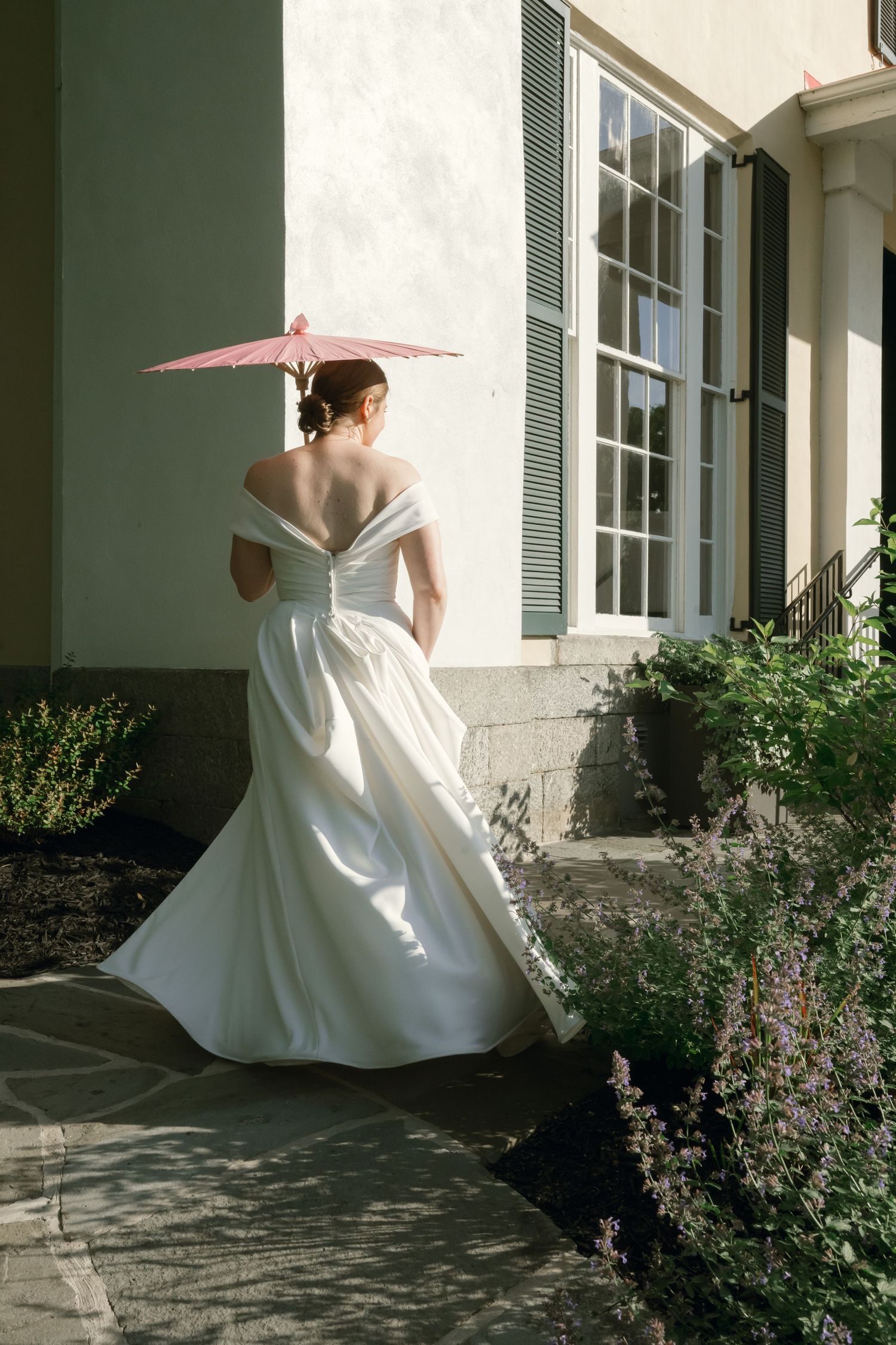 A white wedding dress is showcased against a historic building exterior with green shutters and a pink parasol.