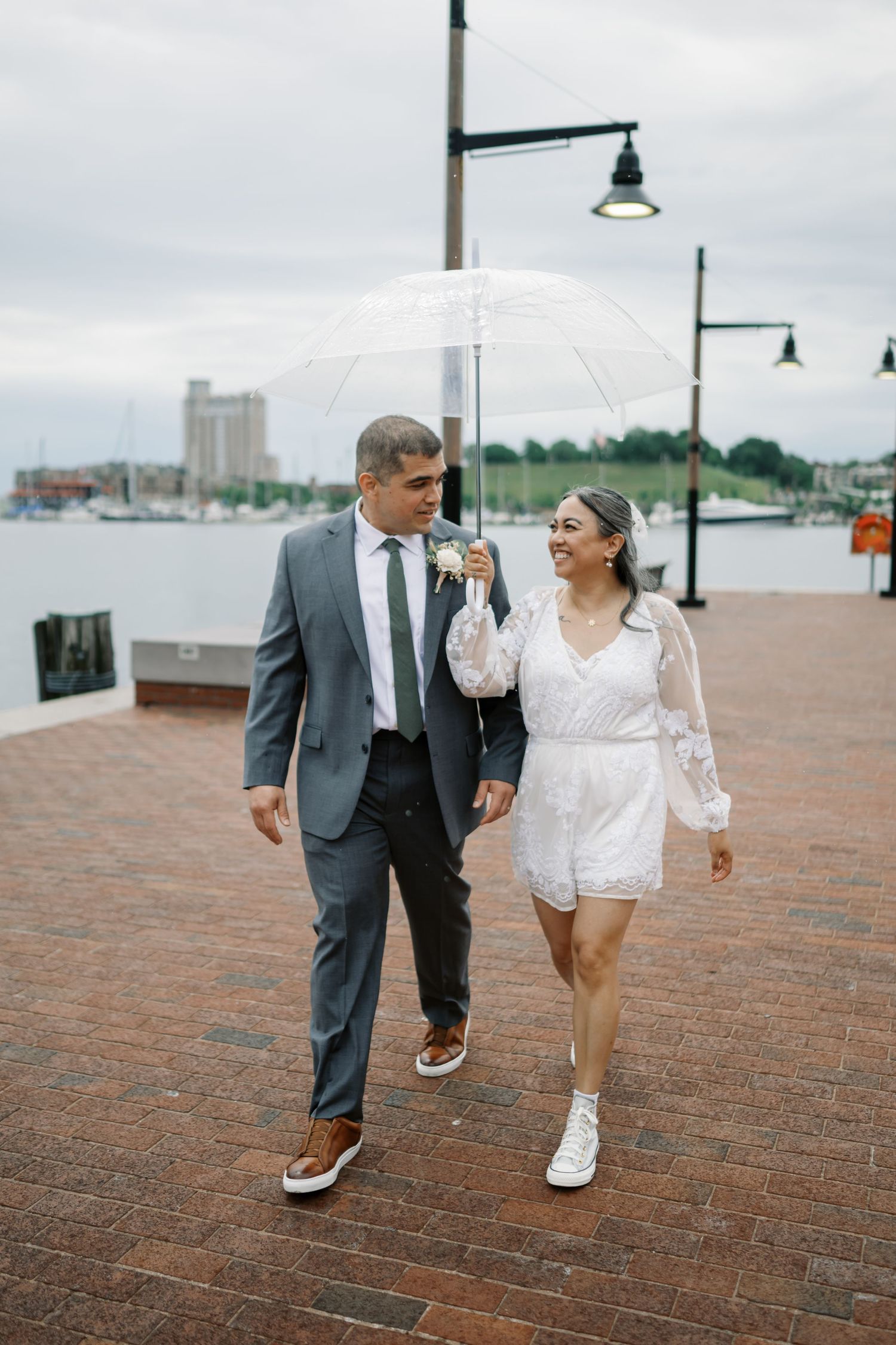 A couple strolls along a brick walkway by the harbor holding a white umbrella.