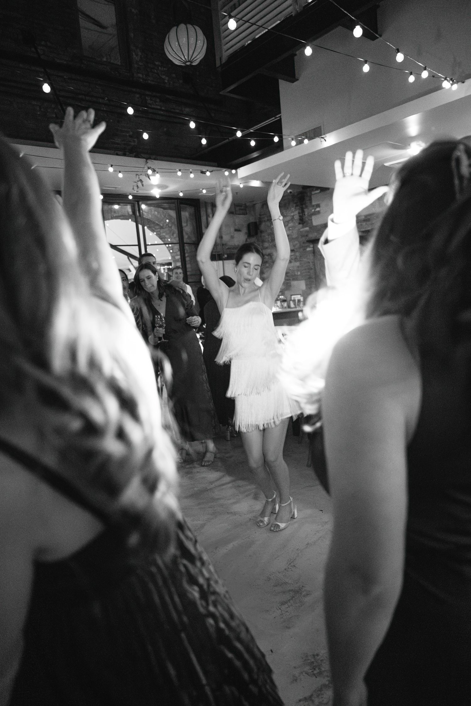 Wedding guests dancing under string lights at an indoor reception venue.