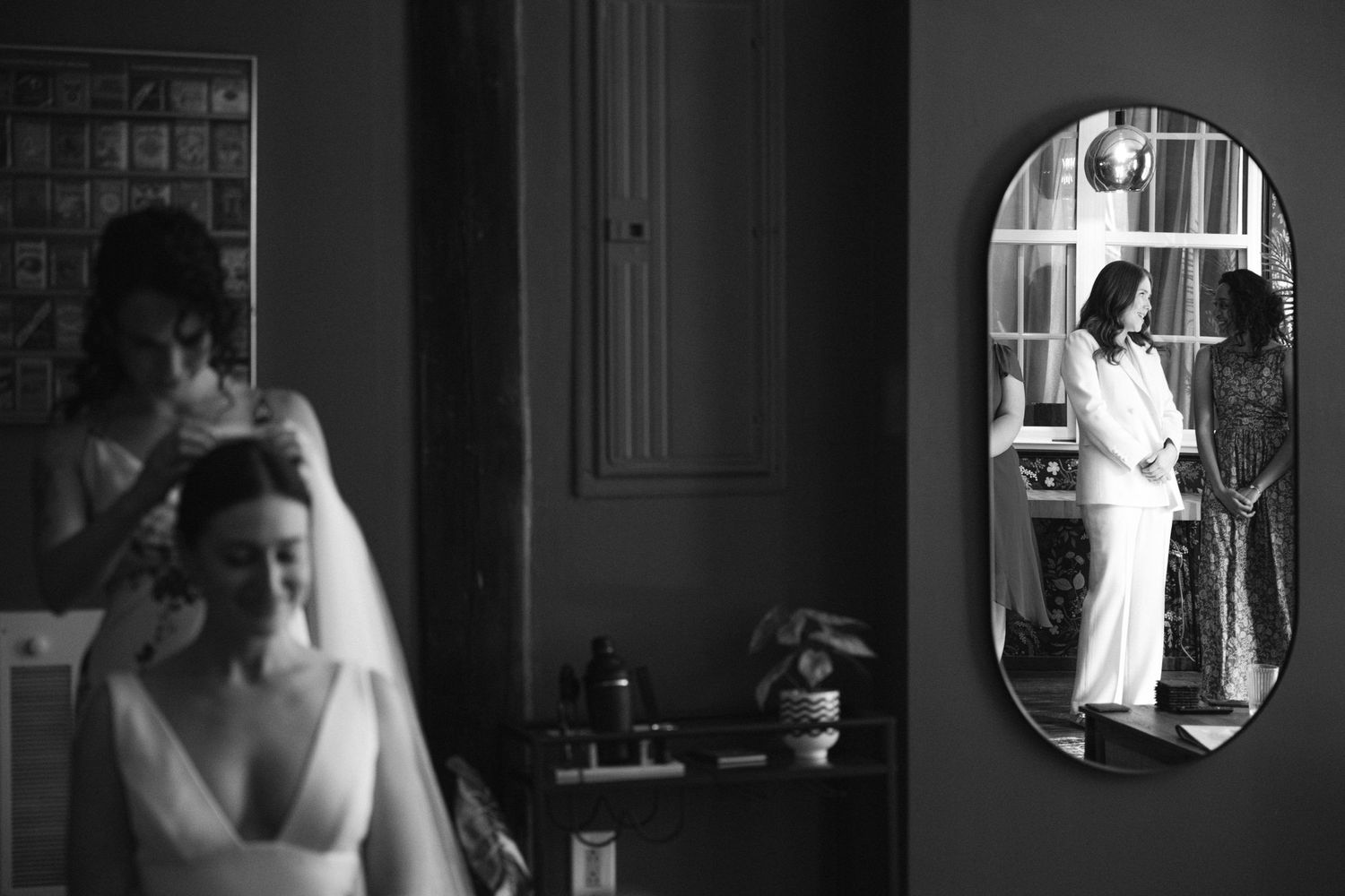 Black and white photo of bride getting veil placed while mirror reflects elegant wedding preparation scene.