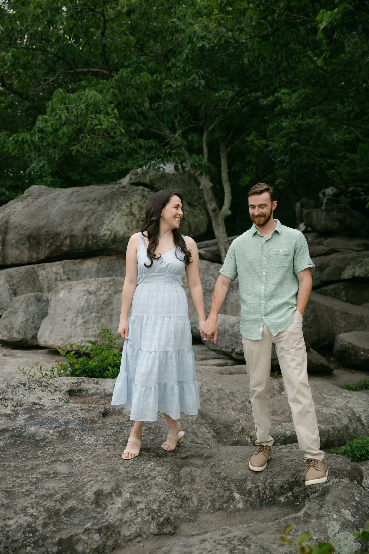 Couple walking hand in hand along rocky terrain with dense forest backdrop in coordinated blue and beige outfits.