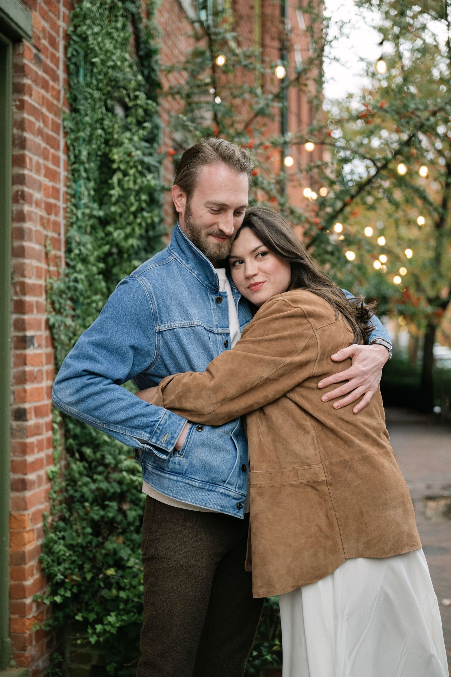 Couple embracing outdoors near brick wall with ivy and string lights, woman in brown suede jacket, man in denim shirt.