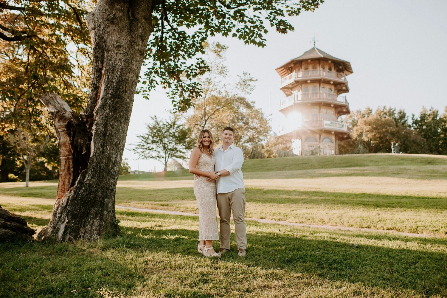 Couple posing together under a large tree in a sunlit park with a historic pagoda tower in the background.