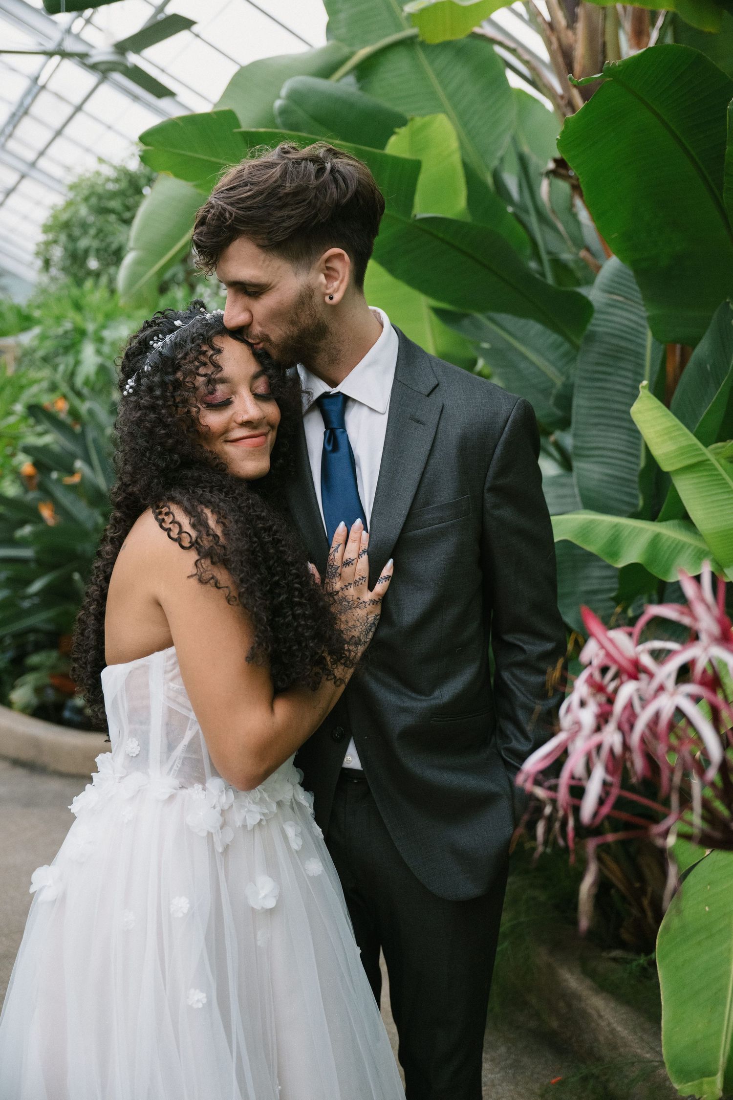 Groom in gray suit kissing bride's forehead as she smiles in a white floral wedding dress in a tropical greenhouse.