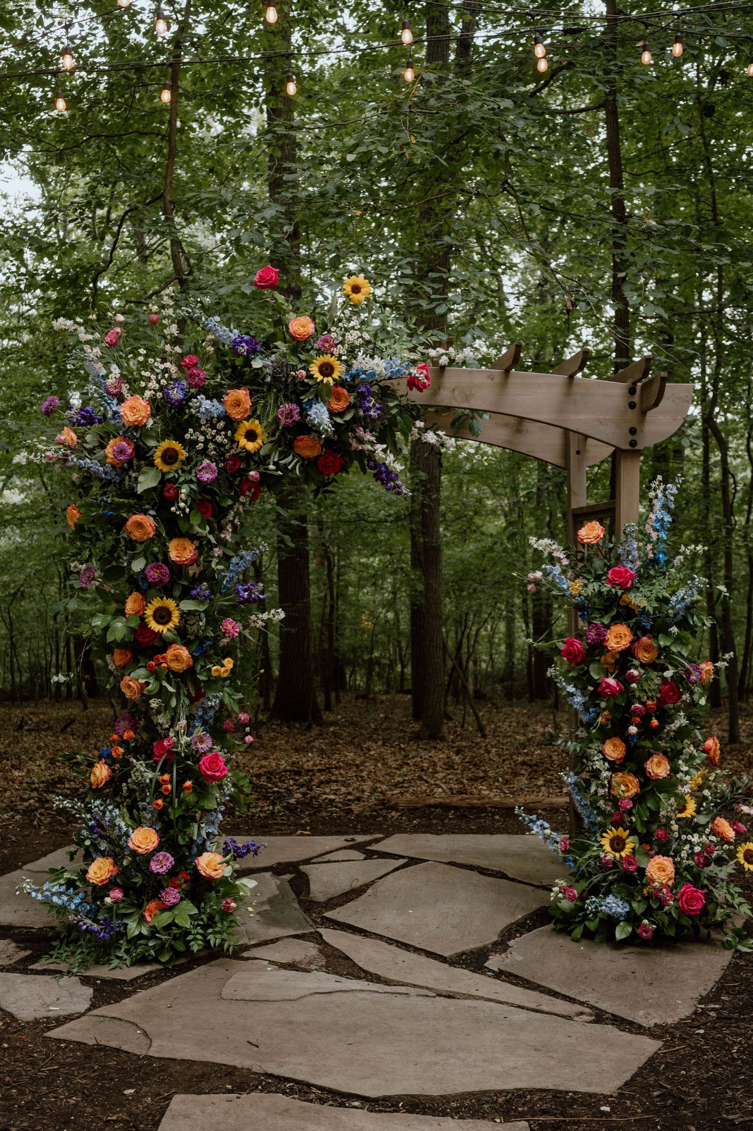 Stunning floral arch with colorful blooms and greenery set on a stone pathway in a forest.