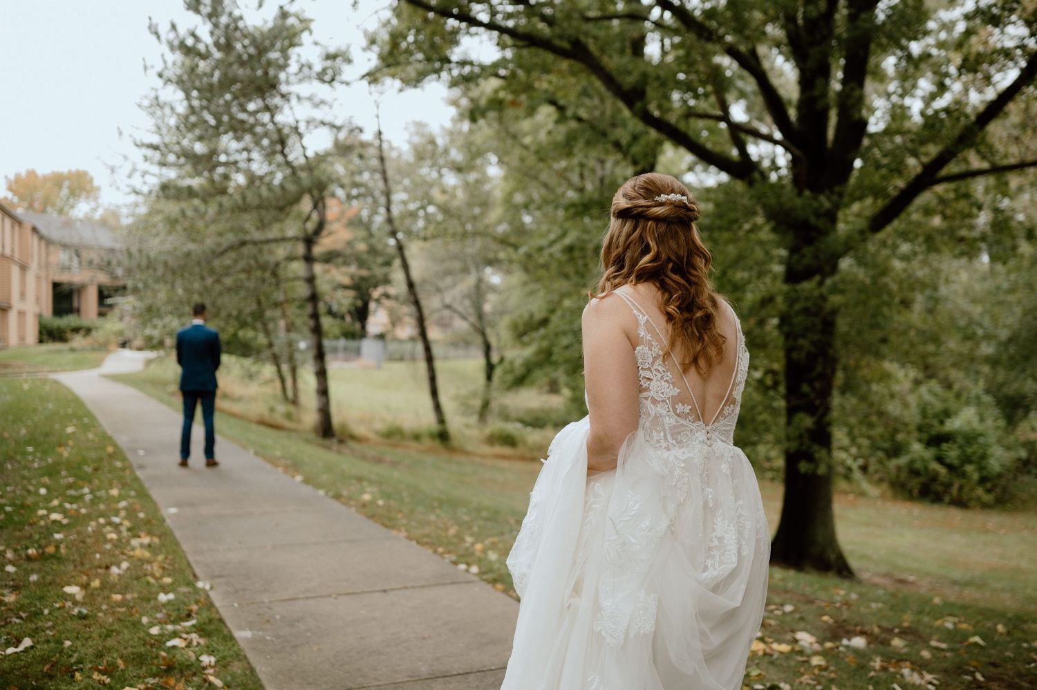 Wedding dress with lace details seen from behind on outdoor path with trees and distant figure.
