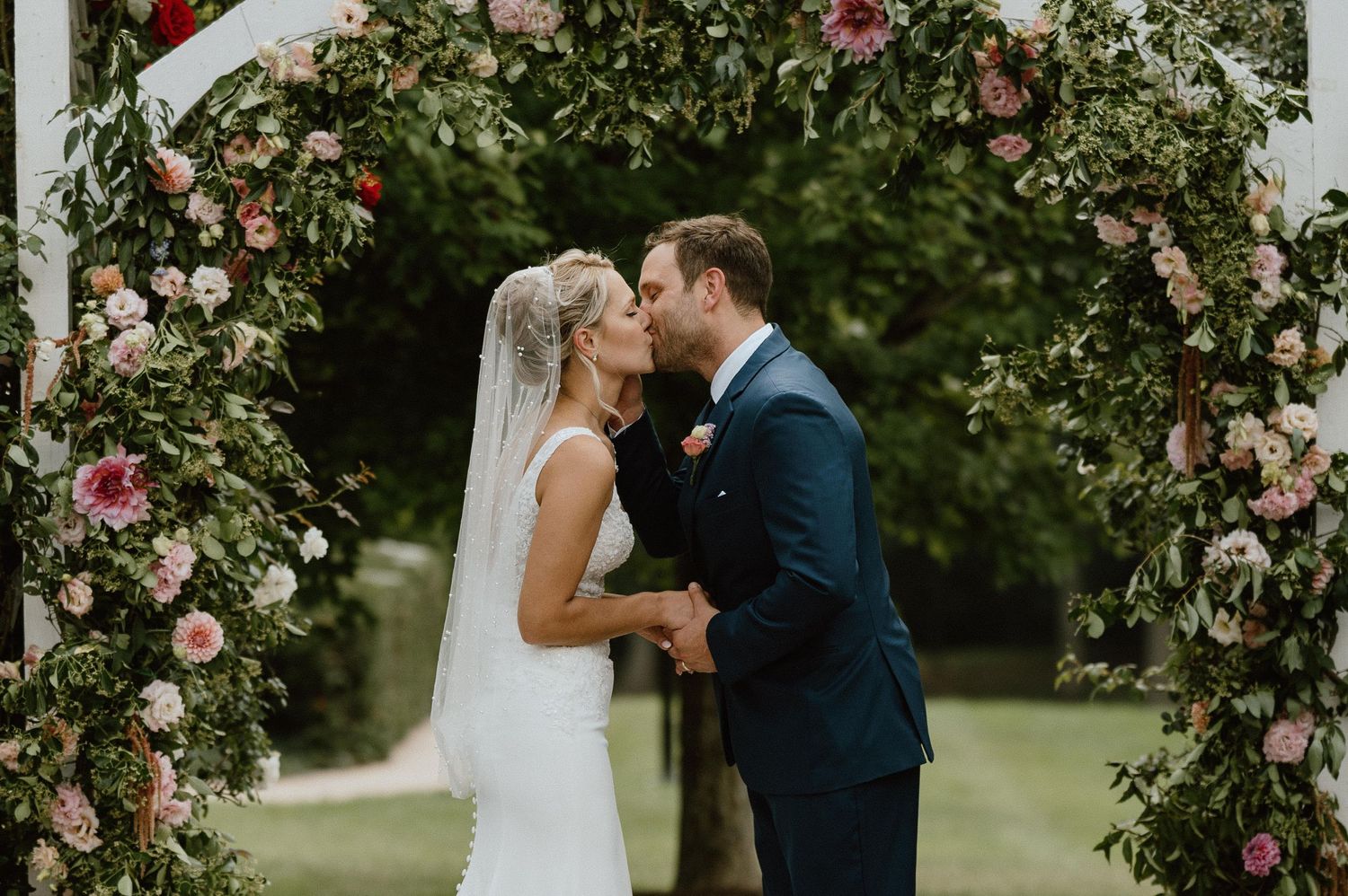 Newlyweds share their first kiss under an arch covered in pink roses and greenery.