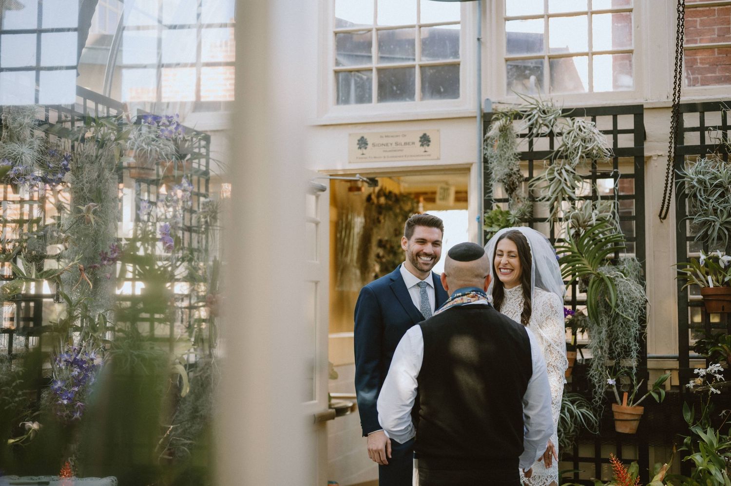 A wedding couple stands in a greenhouse venue with filtered light and indoor plants.