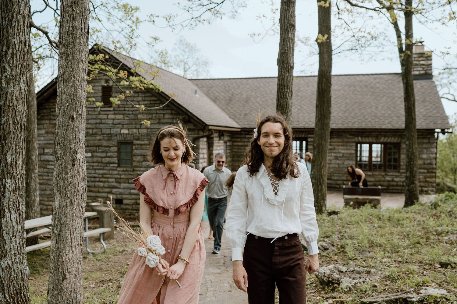 Two people dressed in vintage style clothing walking near a rustic log cabin.