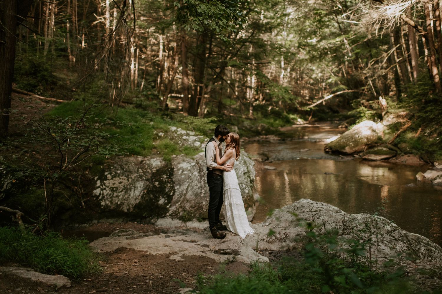 A couple stands together on rocks by a serene forest stream during their wedding.