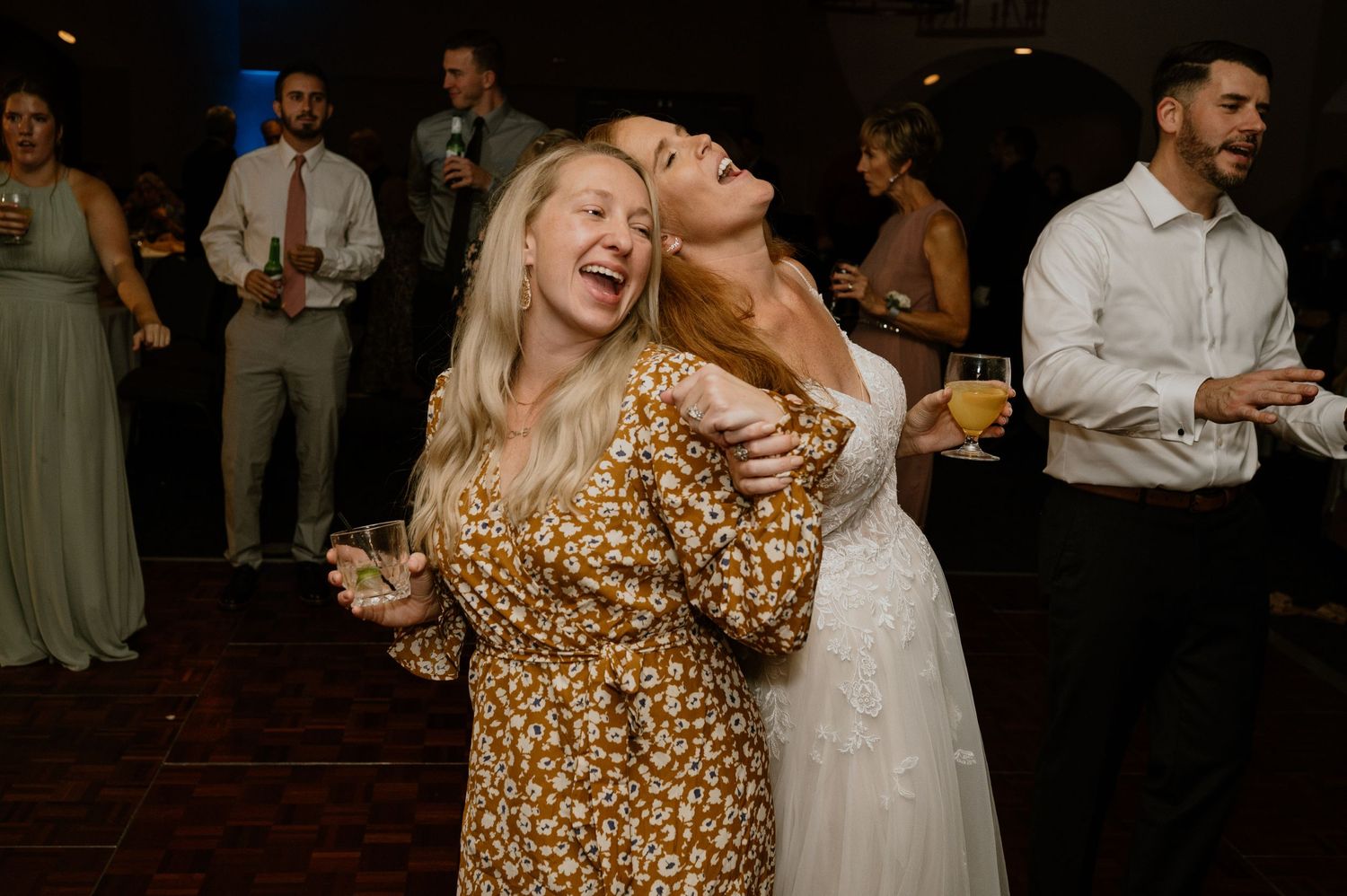 Wedding guests laughing and dancing at indoor reception with dark wood floors.