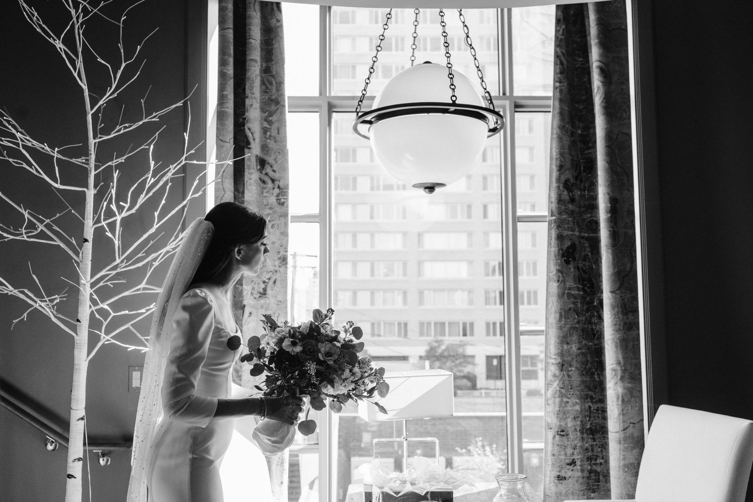 A bride in white holds a bouquet while standing by a window with decorative branches.
