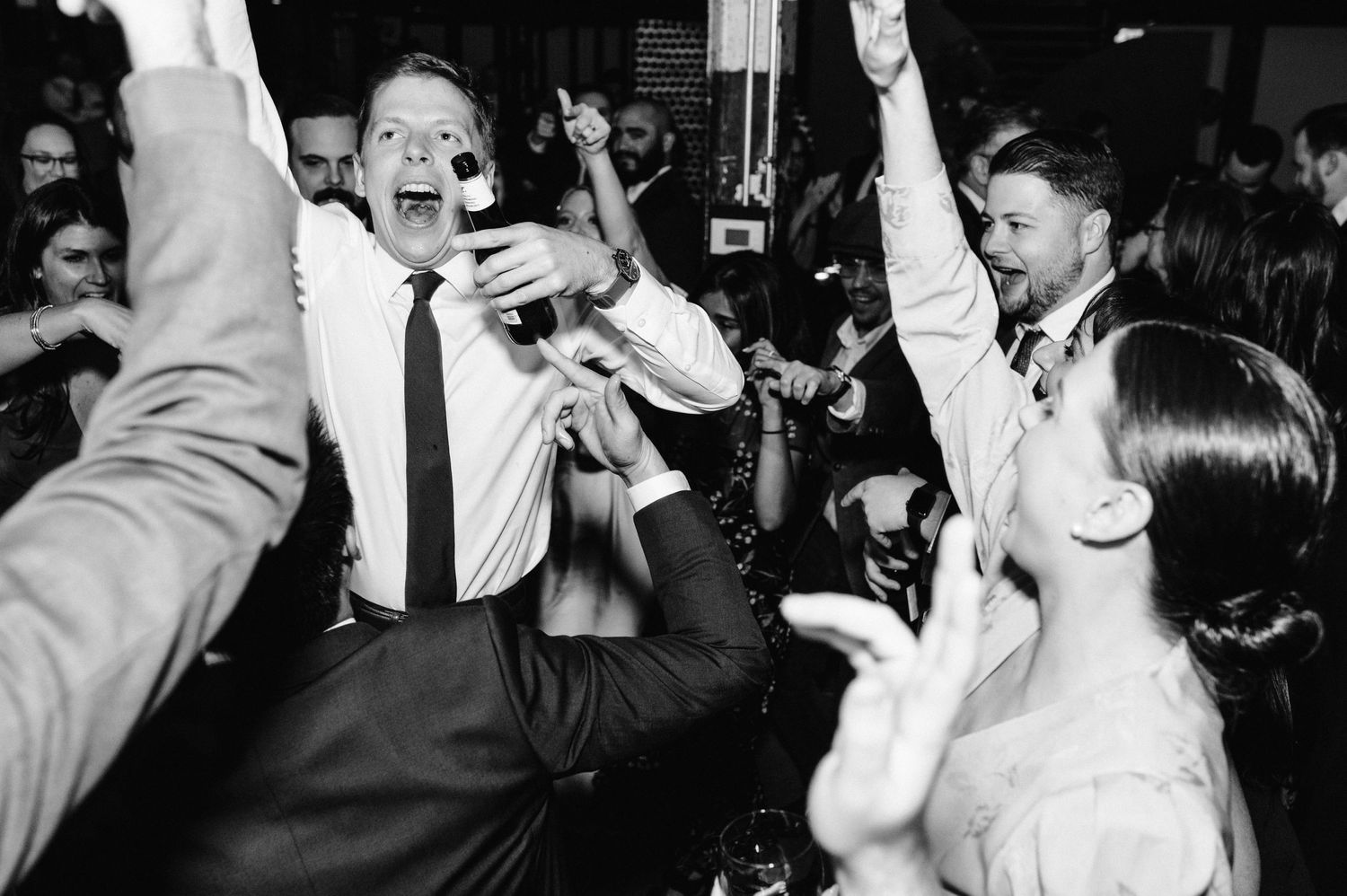 Joyful partygoers dance with their arms raised on a lively dance floor in black and white.